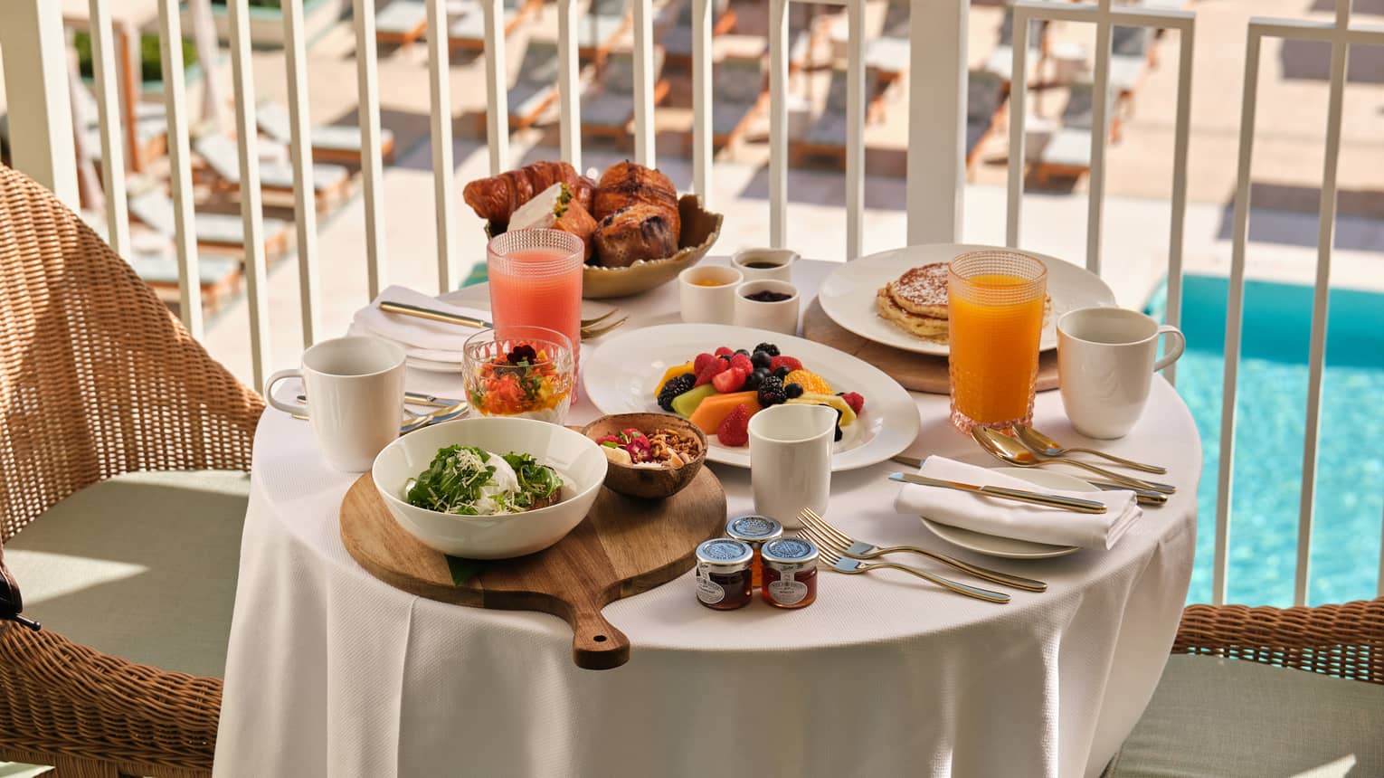 Three plates of breakfast food, a bowl of pastries, two coffee mugs and two glasses of juice set on a small round table on a hotel balcony overlooking a pool