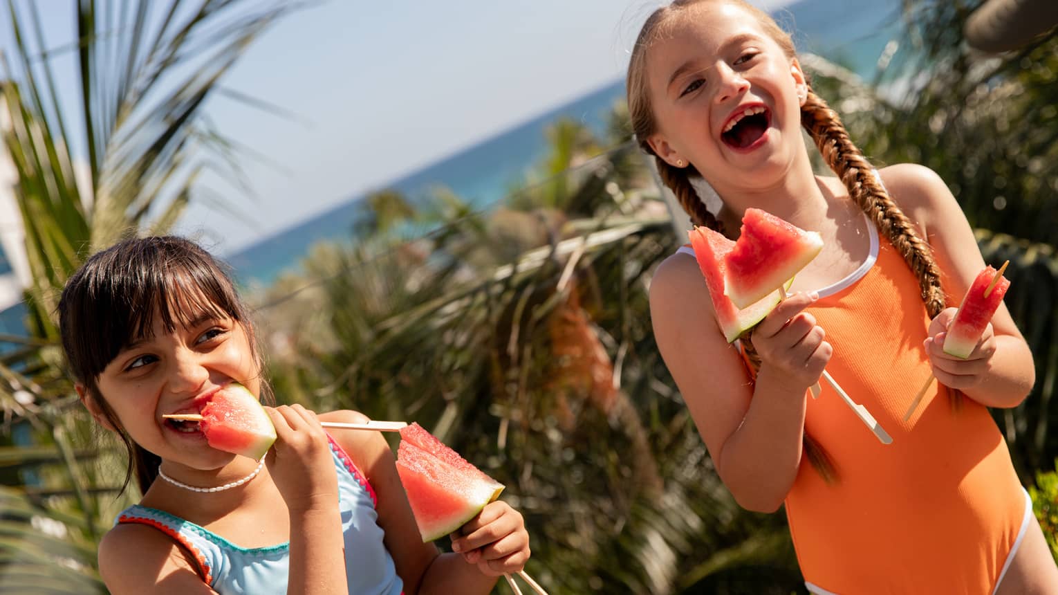 Two smiling children wearing bright-coloured bathing suits stand near palm trees eating slices of watermelon served on sticks