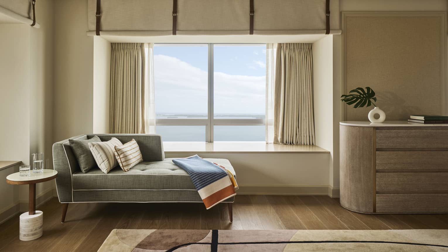 A chaise longue with blanket is positioned next to a picture window with view of the sea, in a hotel suite
