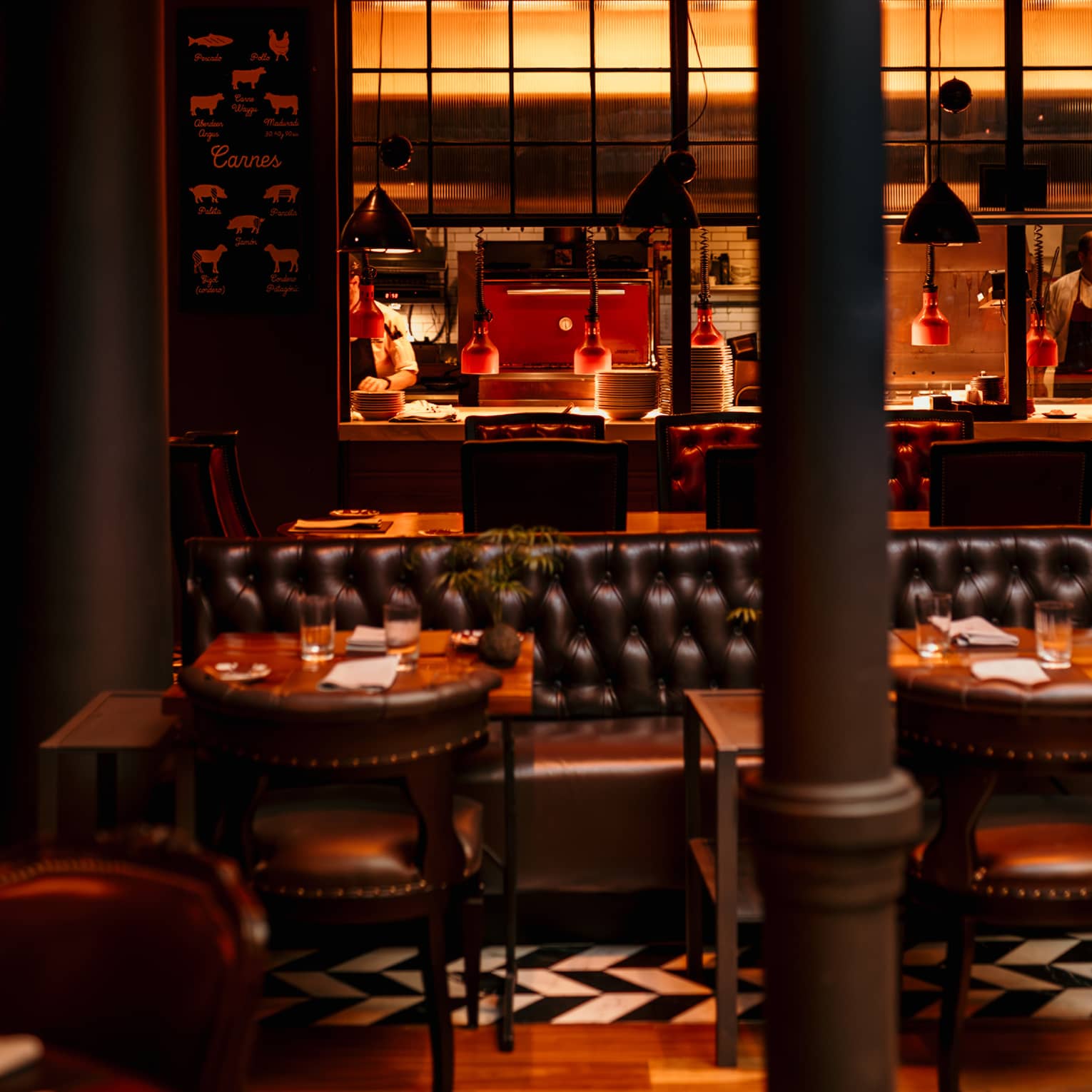 Dimly lit restaurant dining room with a tufted leather banquette, leather chairs and an open kitchen