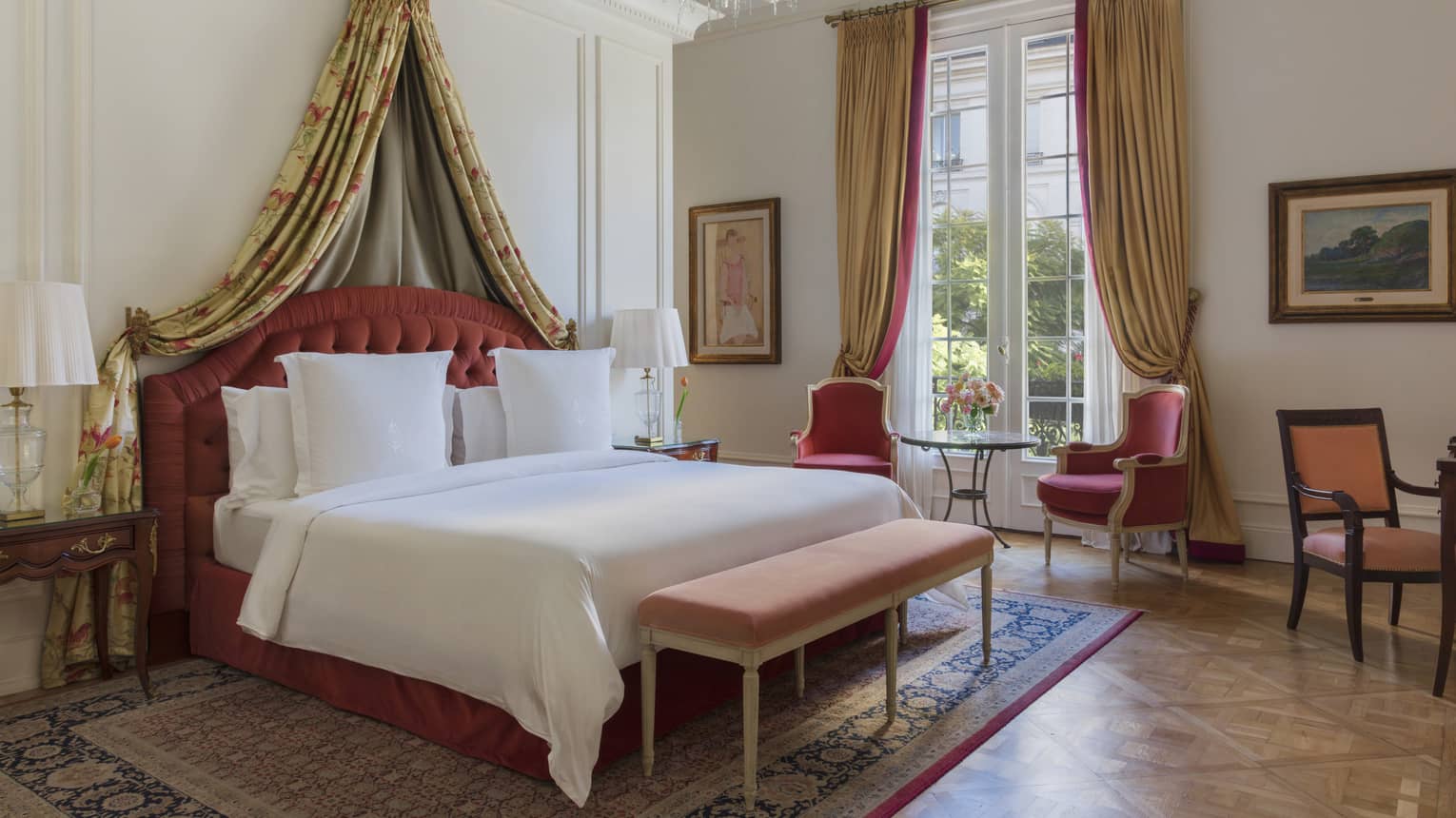 Lavish hotel suite bedroom, featuring a king bed with draped canopy and red armchairs by a window