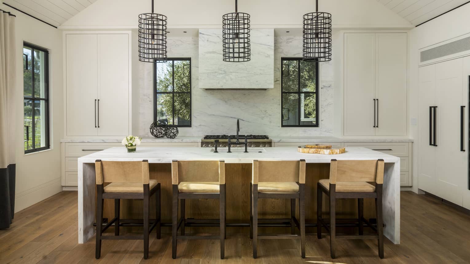 Luxury resort kitchen with marble island, four beige barstools, black pendant lights, large white cabinets, stove and three windows overlooking greenery