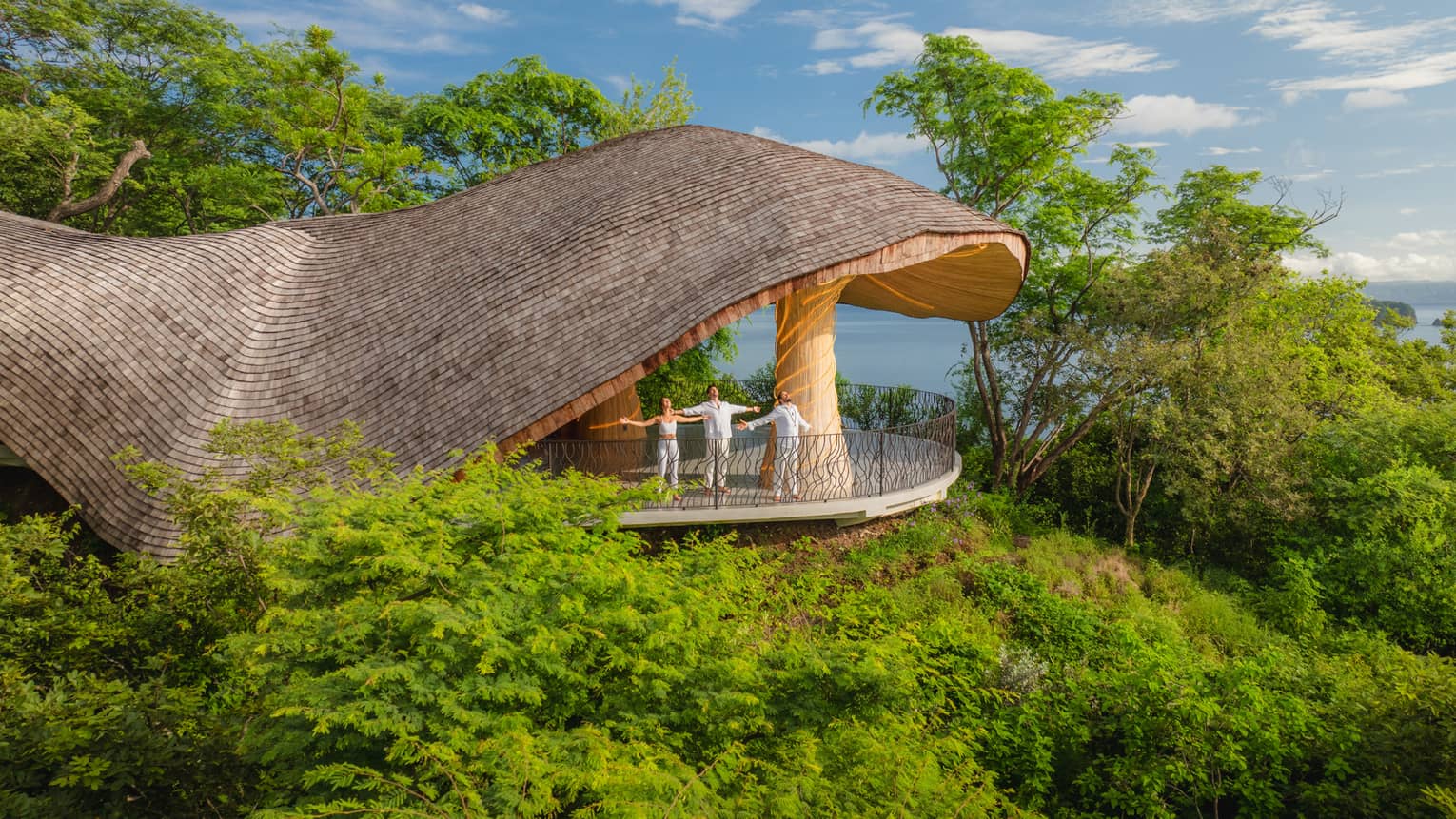 Threee people stand with arms stretched open at the edge of a cliffside yoga pavillion overlooking a green landscape with the ocean in the background