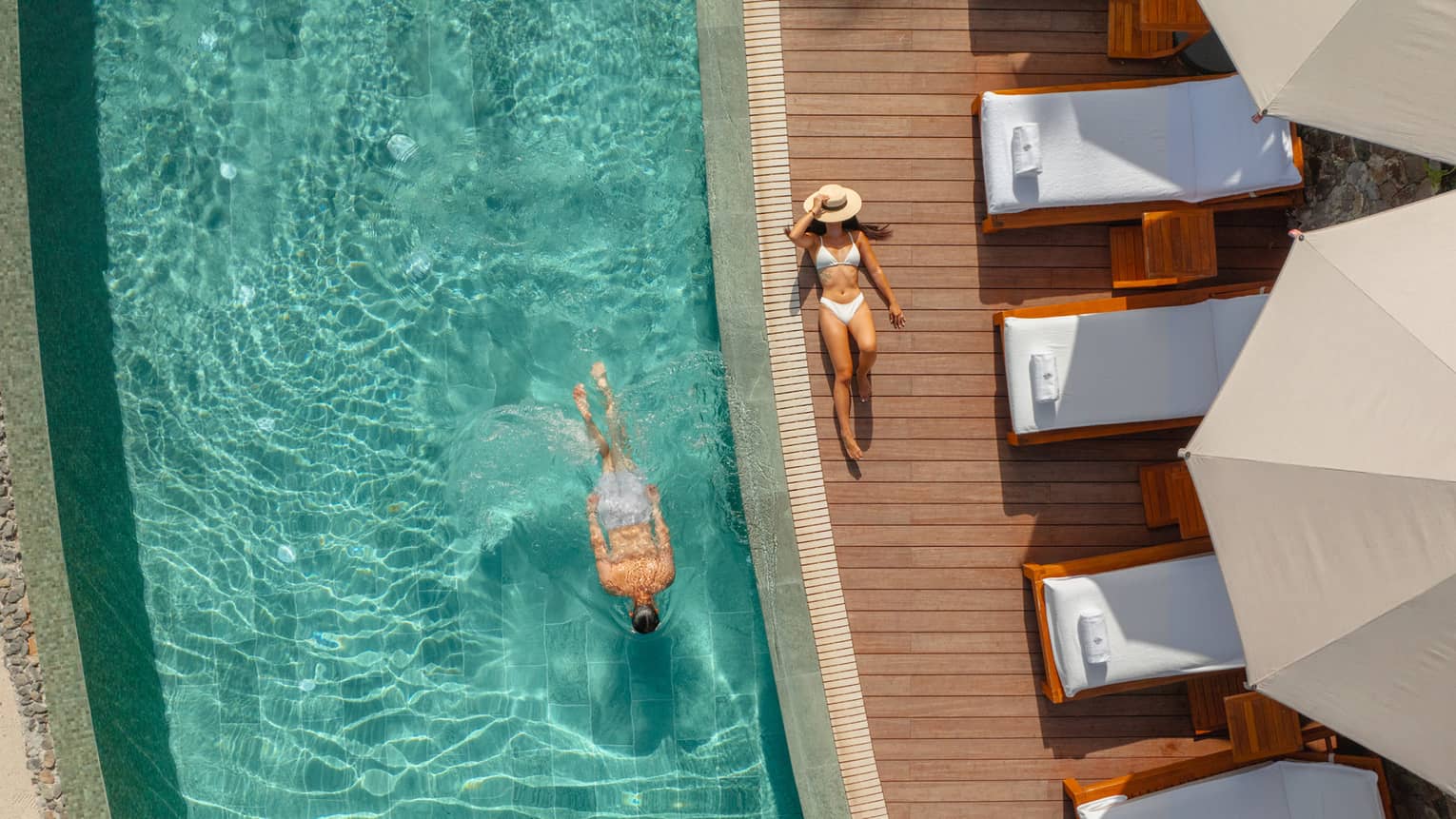 Aerial view of a man swimming in a pool with a woman in a white bikini lying next to the water on the floor of the pool deck