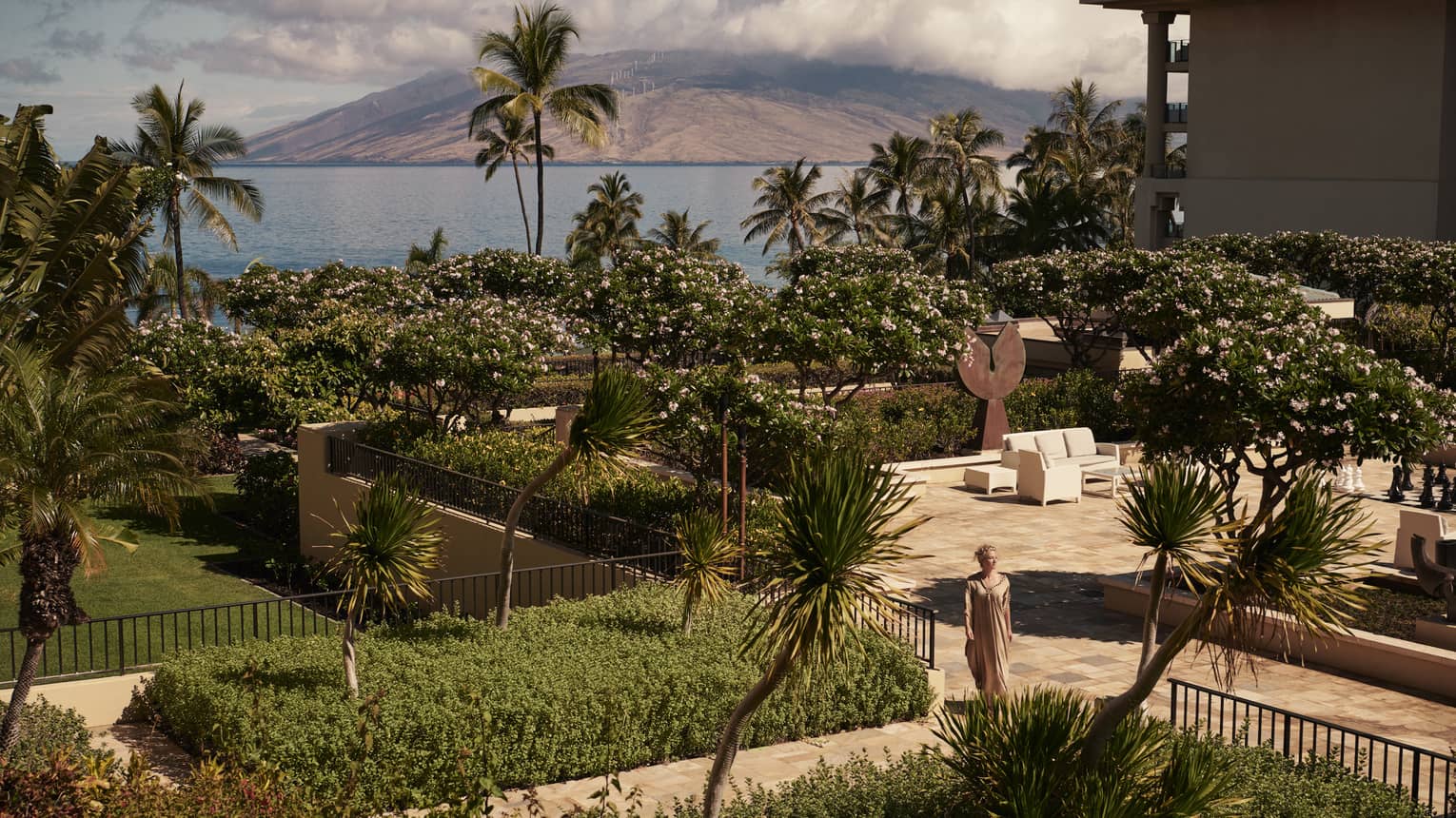 Woman walks through gardens on resort grounds in Maui, ocean in background