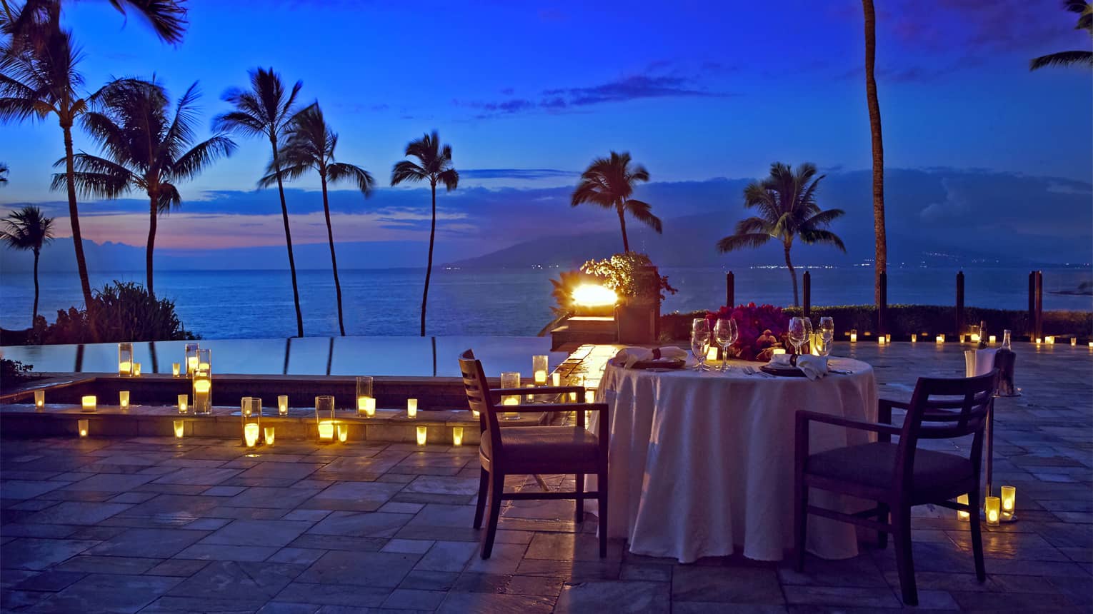 Romantic oceanfront dinner table setup at dusk with candles and palm trees