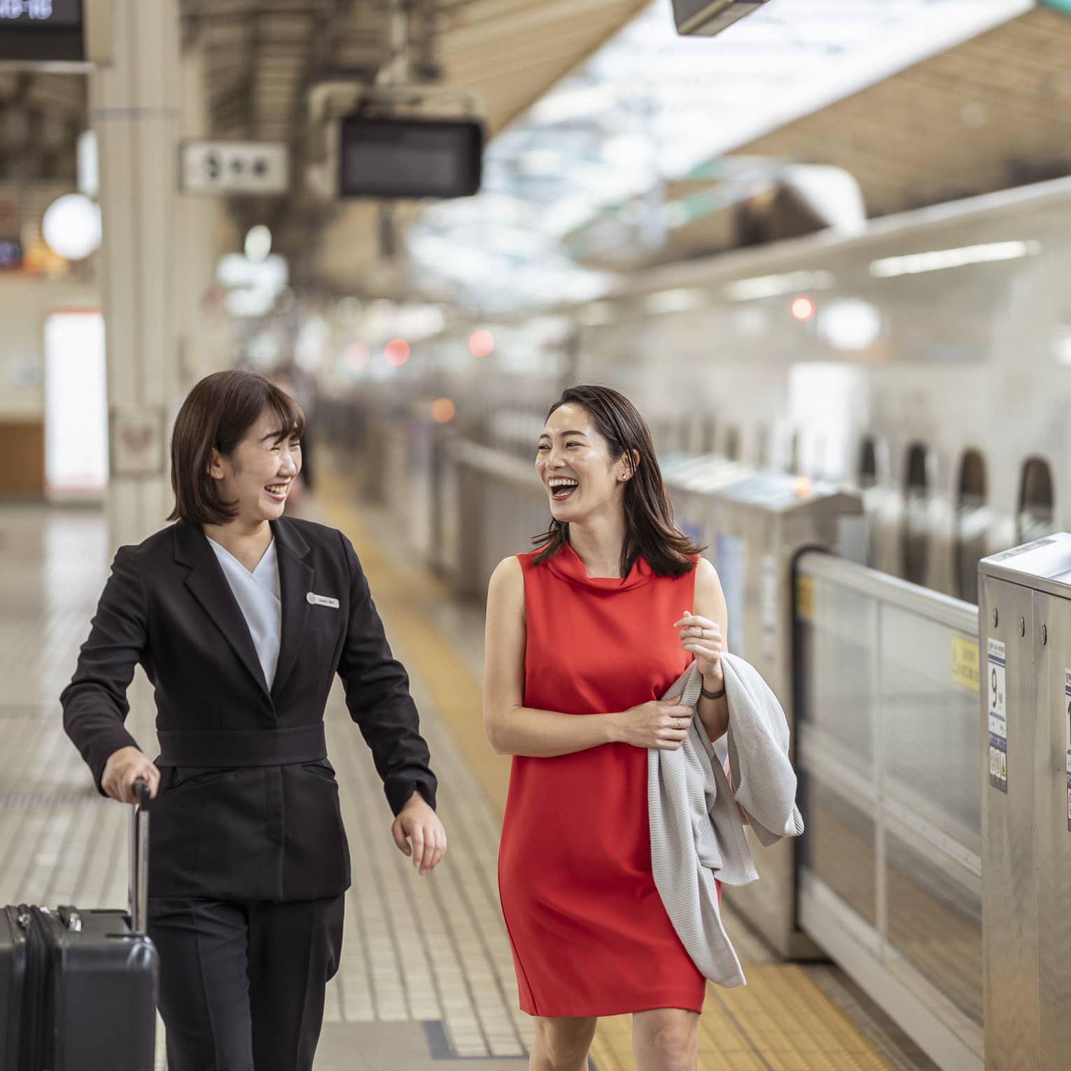 A Four Seasons employee holds a traveller's suitcase; laughing, they walk along the platform beside an N700 high-speed train.