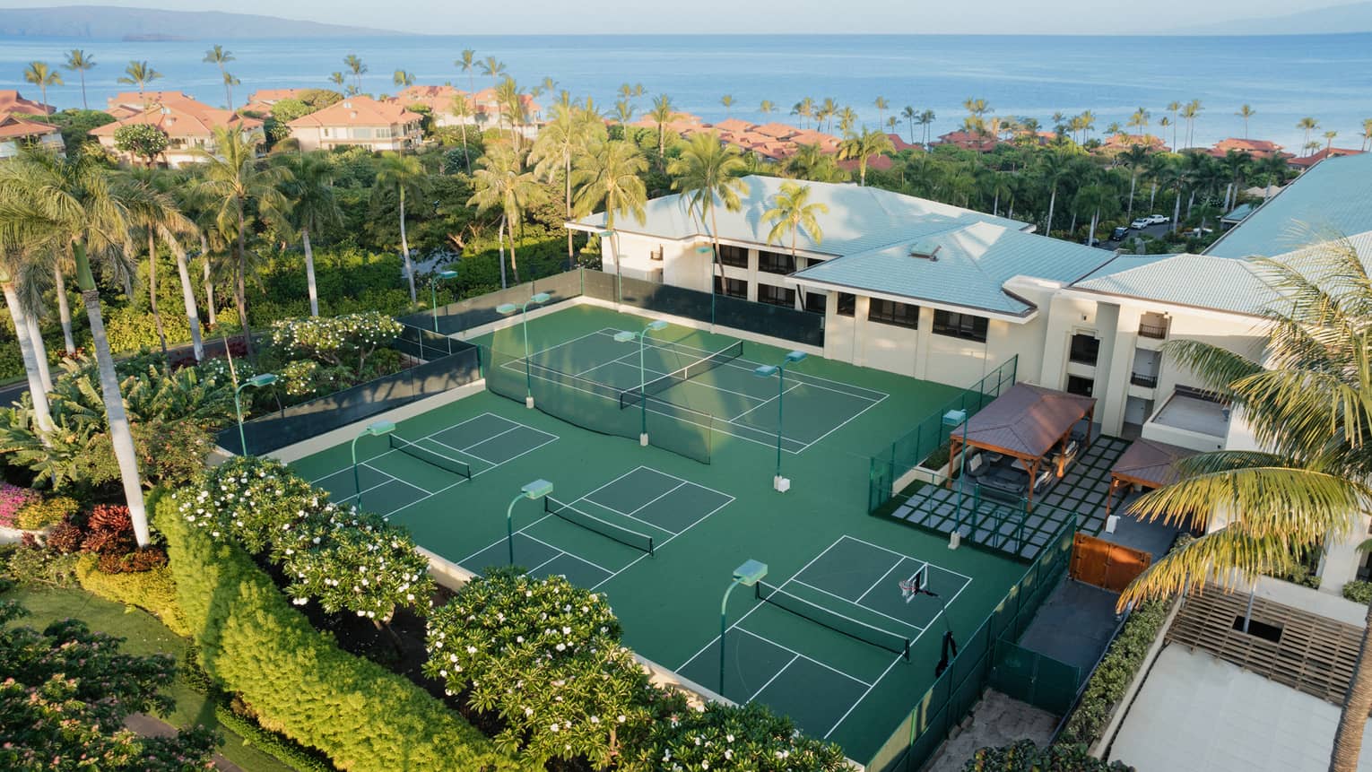 An aerial view of a group of pickleball courts adjacent to a white building, all surrounded by greenery and palm trees, with the ocean in the background