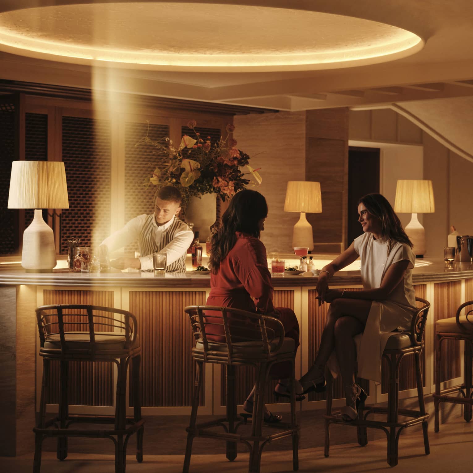 Two smiling guests sit at a warmly lit circular bar at night as a bartender prepares cocktails in a stream of golden light.