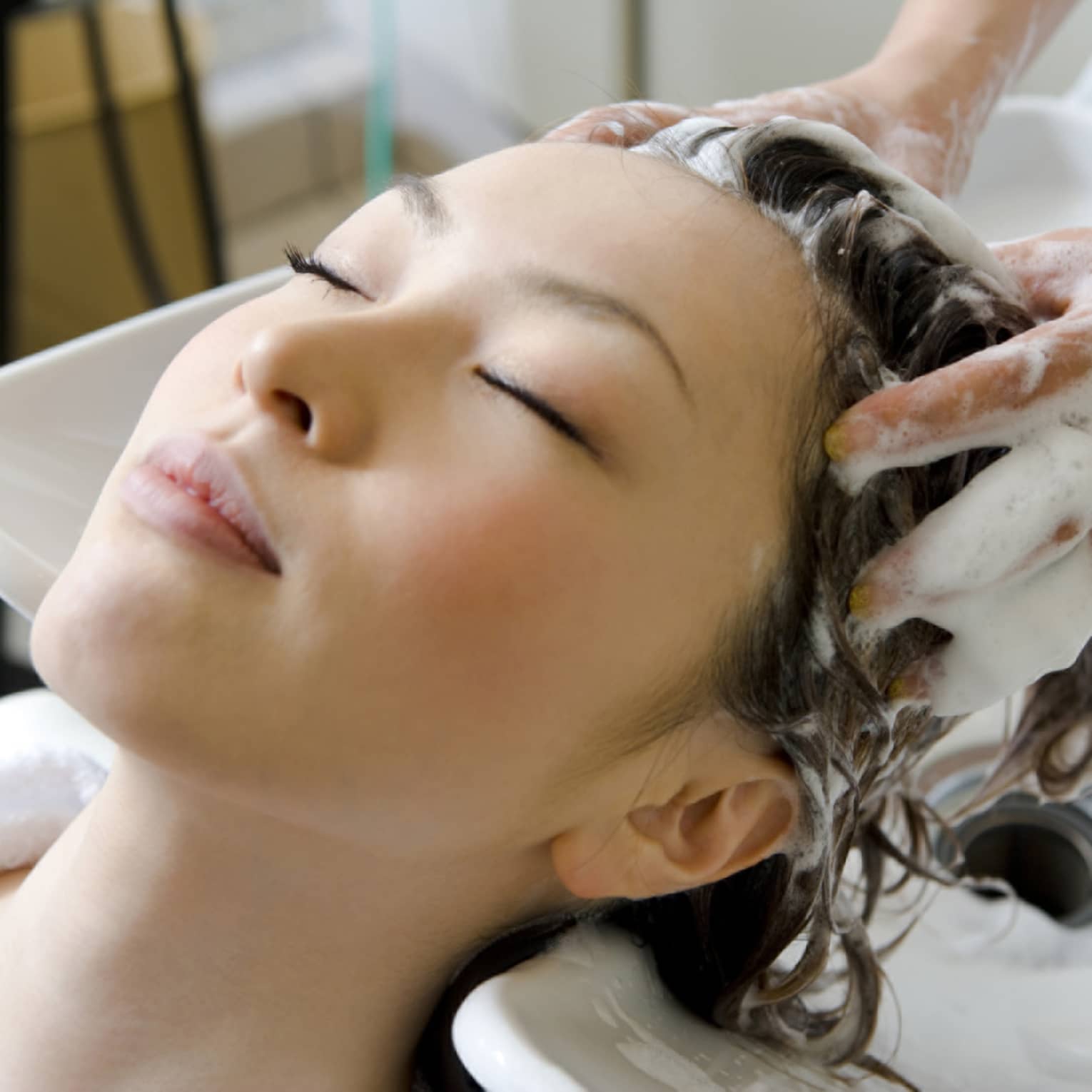 Woman closes eyes and leans back against sink as hands massage shampoo into her scalp