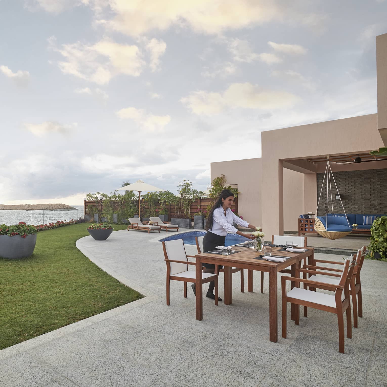 A chef seasons meat on a barbecue near the water’s edge while a server arranges flowers at a table on a patio near a pool.