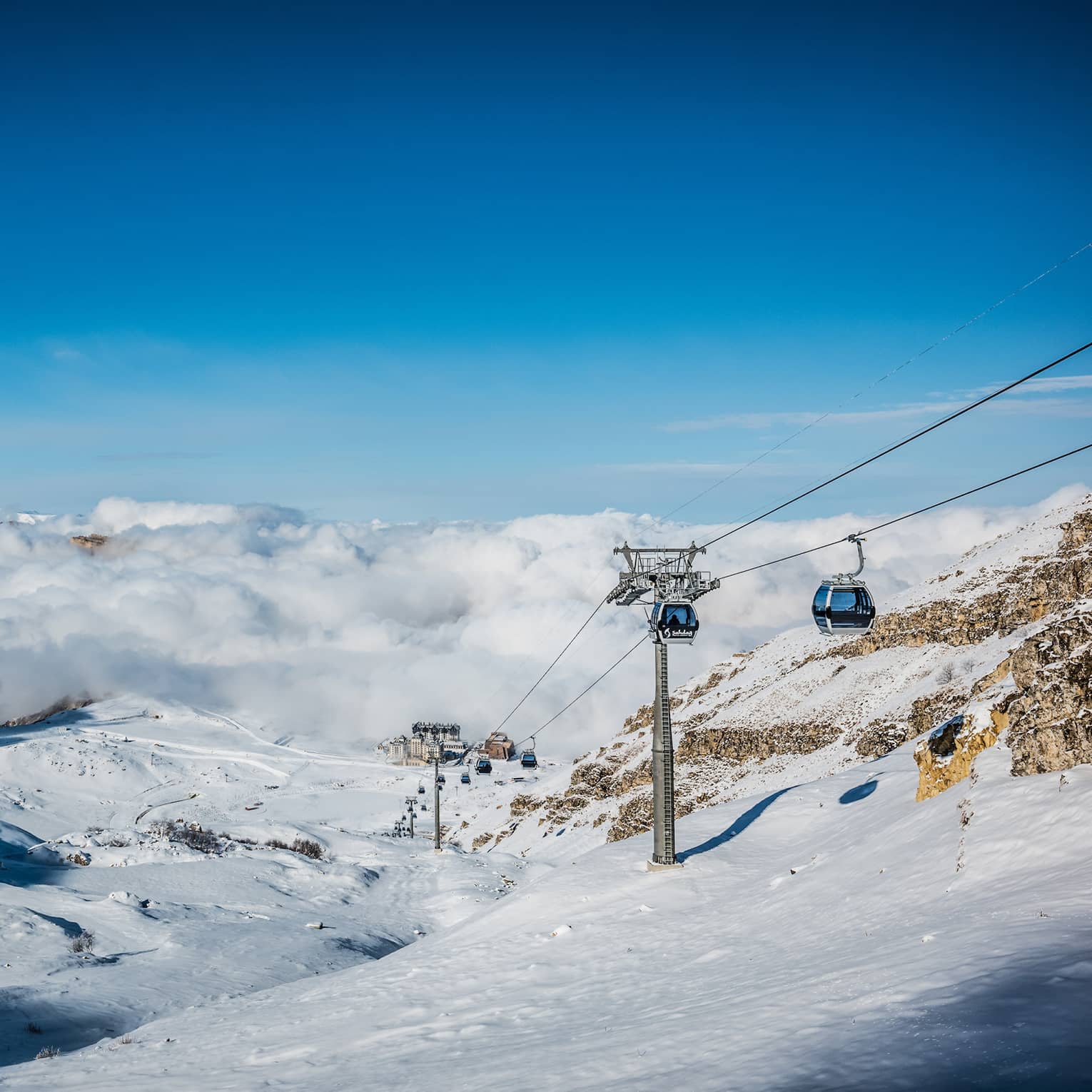 Ski lift cars pass over snowy mountainous landscape with blue sky in background