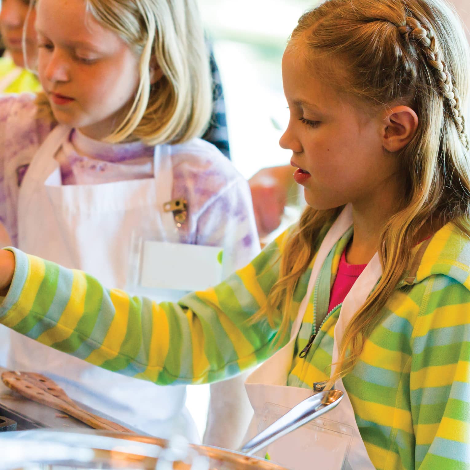 Two young girls in aprons stir pan on stove during Thai cooking class