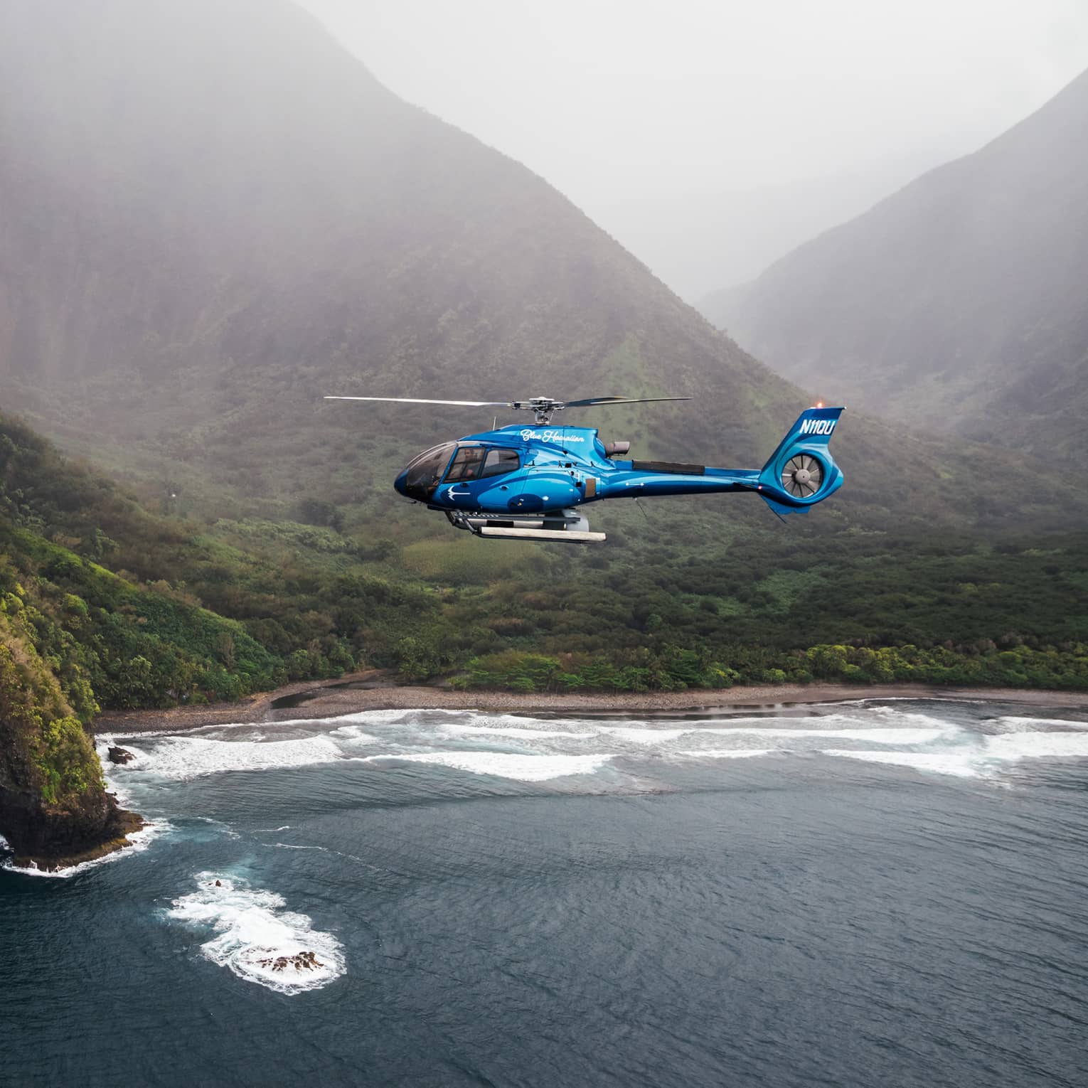 A bright blue helicopter flying above dark blue ocean waters with a verdant, mountainous coastline and misty skies in the background