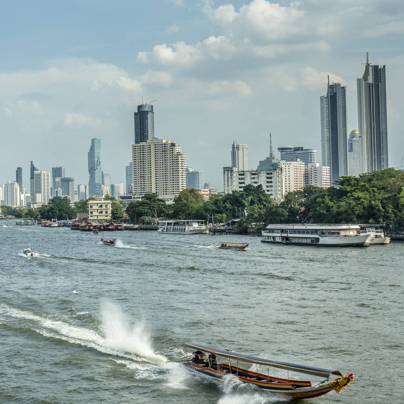 Longtail boat heading into foreground away from Bangkok skyline, leaving trail behind and surrounded by tall, splashing wake.