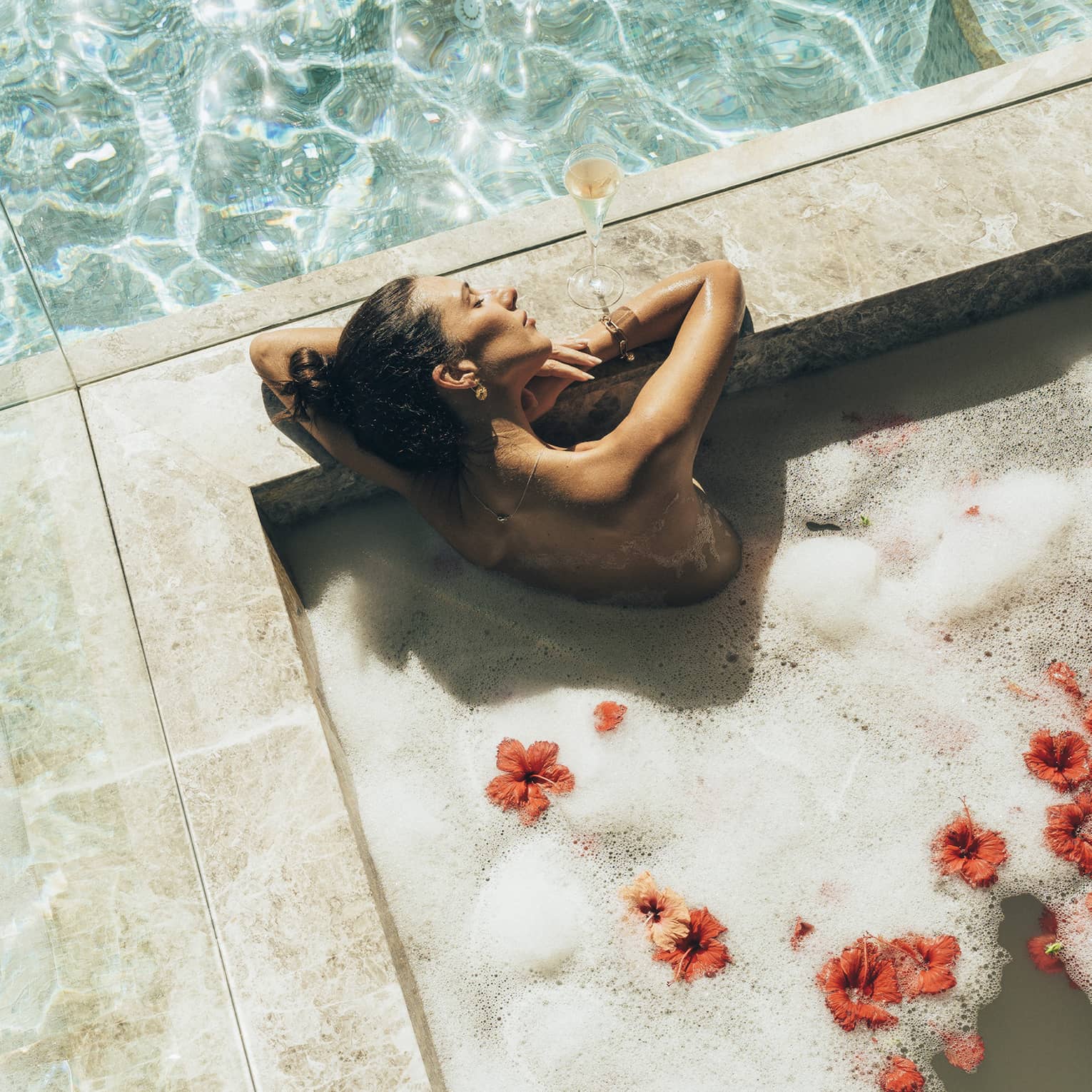Surrounded by mirrored walls, a bather relaxes, eyes closed, in a marble tub of frothy water and red hibiscus blossoms.