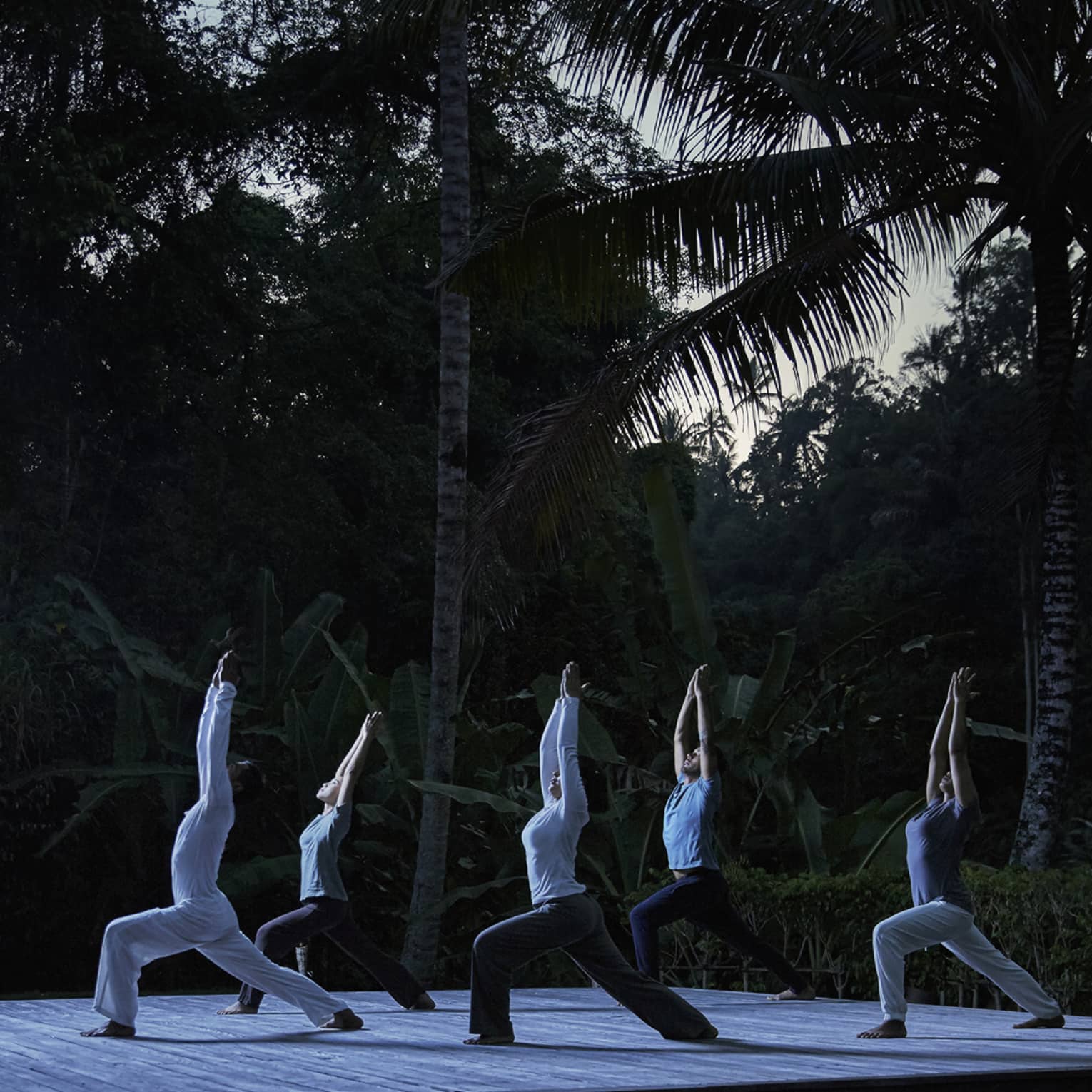 Five people kneel in yoga pose with arms above head on wood platform in forest at night