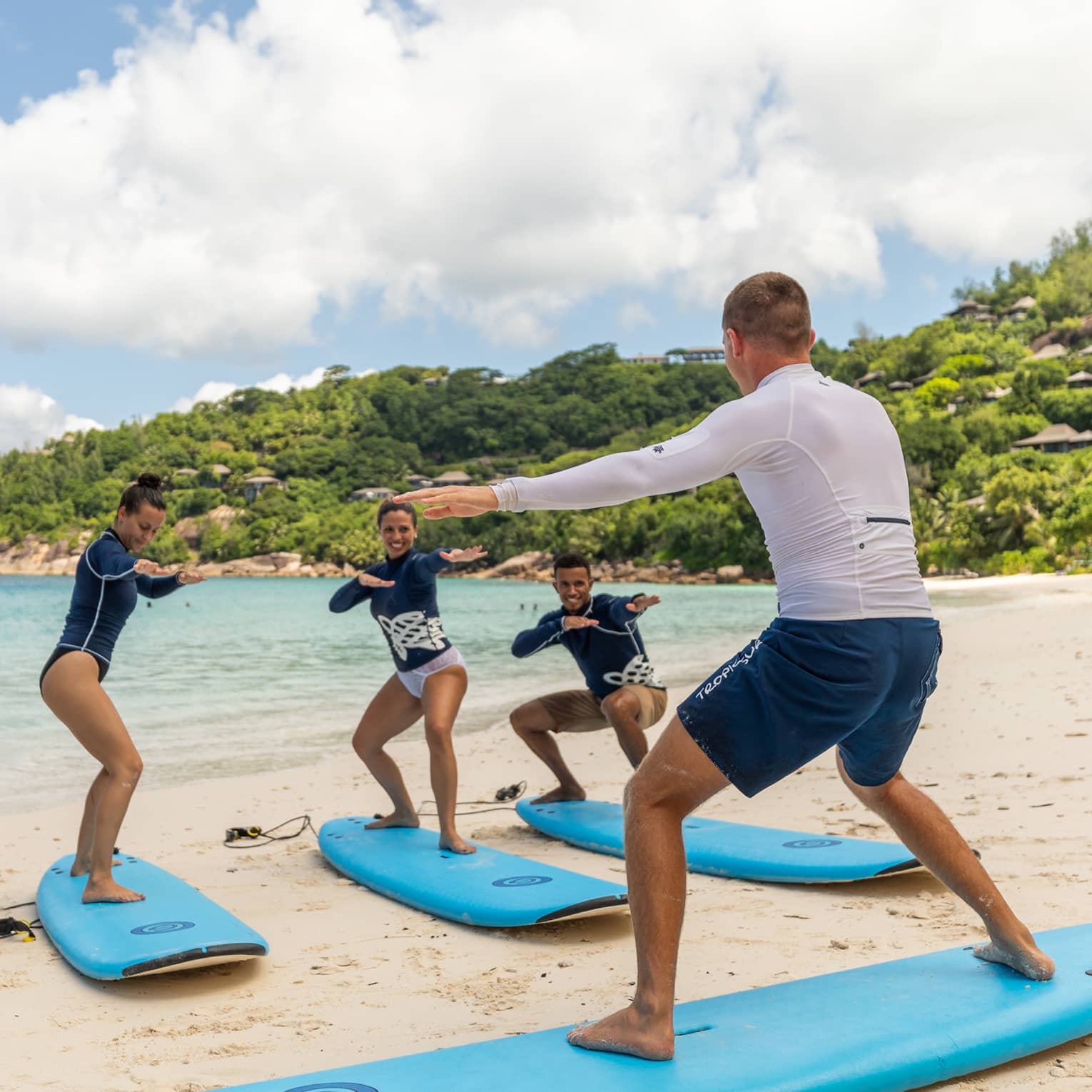 Surfing instructor balances on surfboard on white sand beach in front of row of surfers