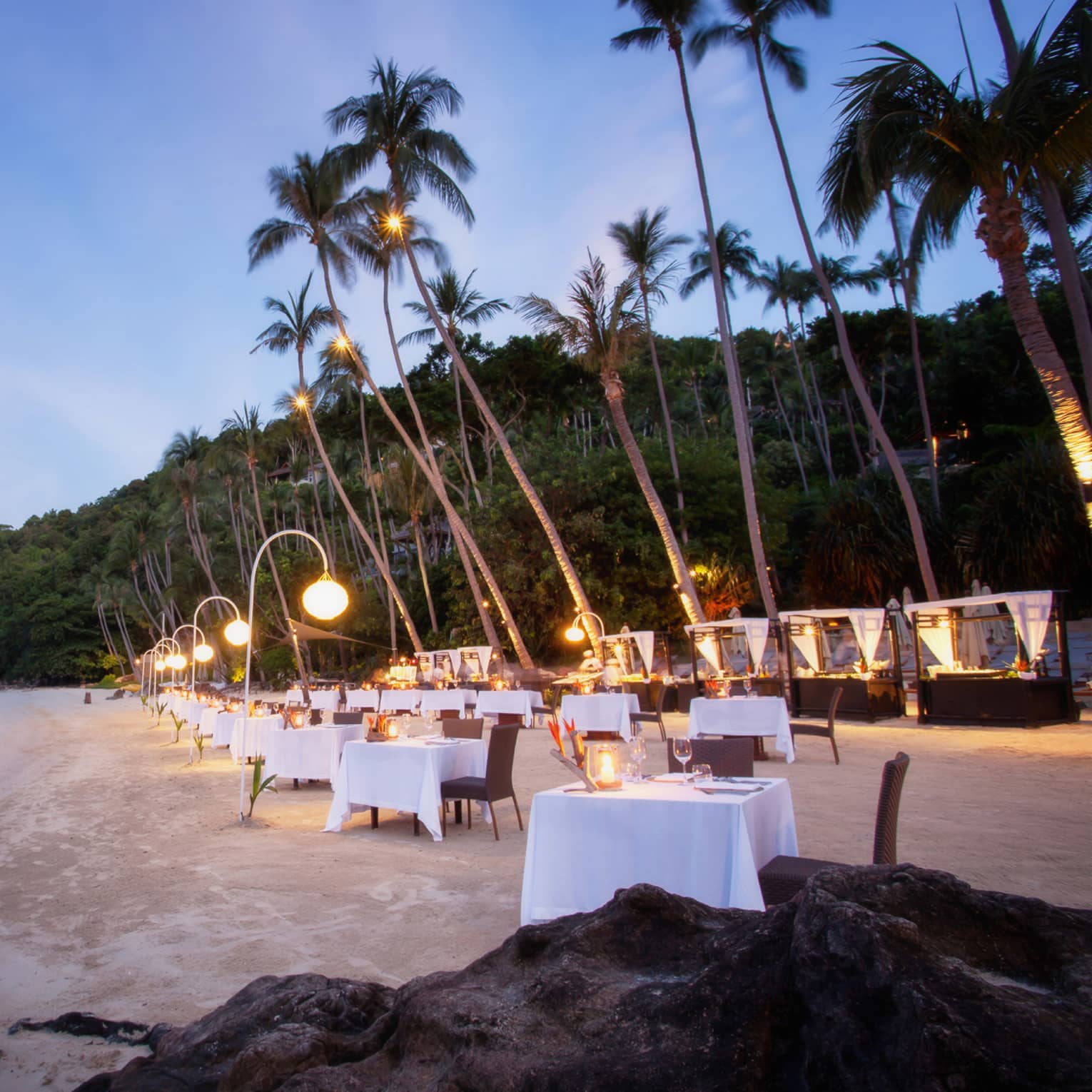 Elegant beachside dining setup at dusk with white-clothed tables, glowing lanterns and tall palm trees