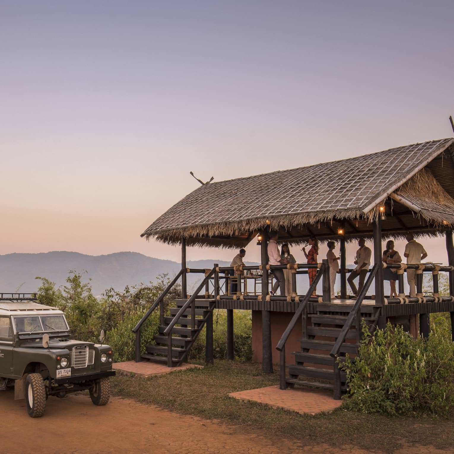 People gather on a raised thatched deck near a campfire and off-road vehicle at sunset.