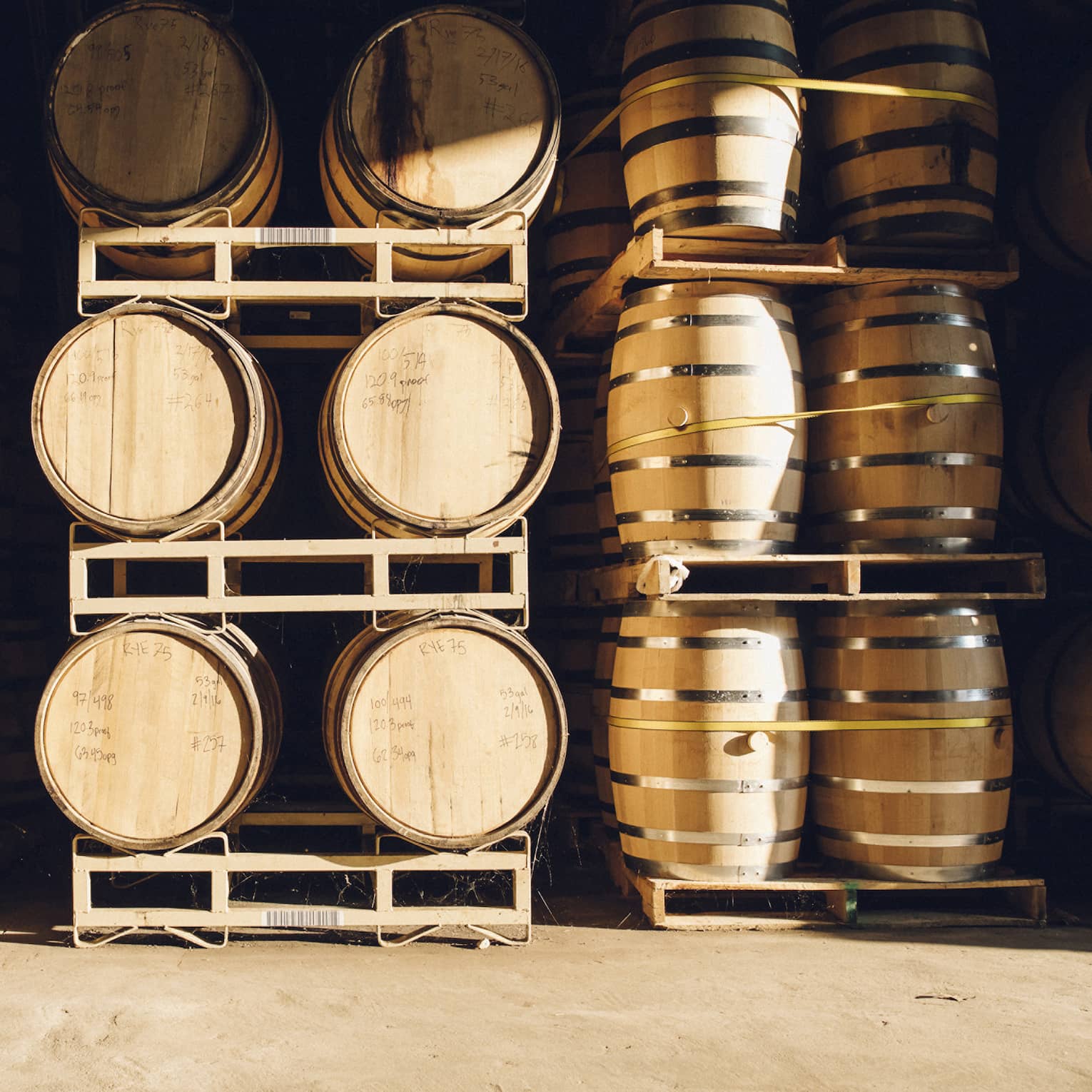 Several wooden barrels lined up on racks in a distillery with information about what is in each barrel written on top.