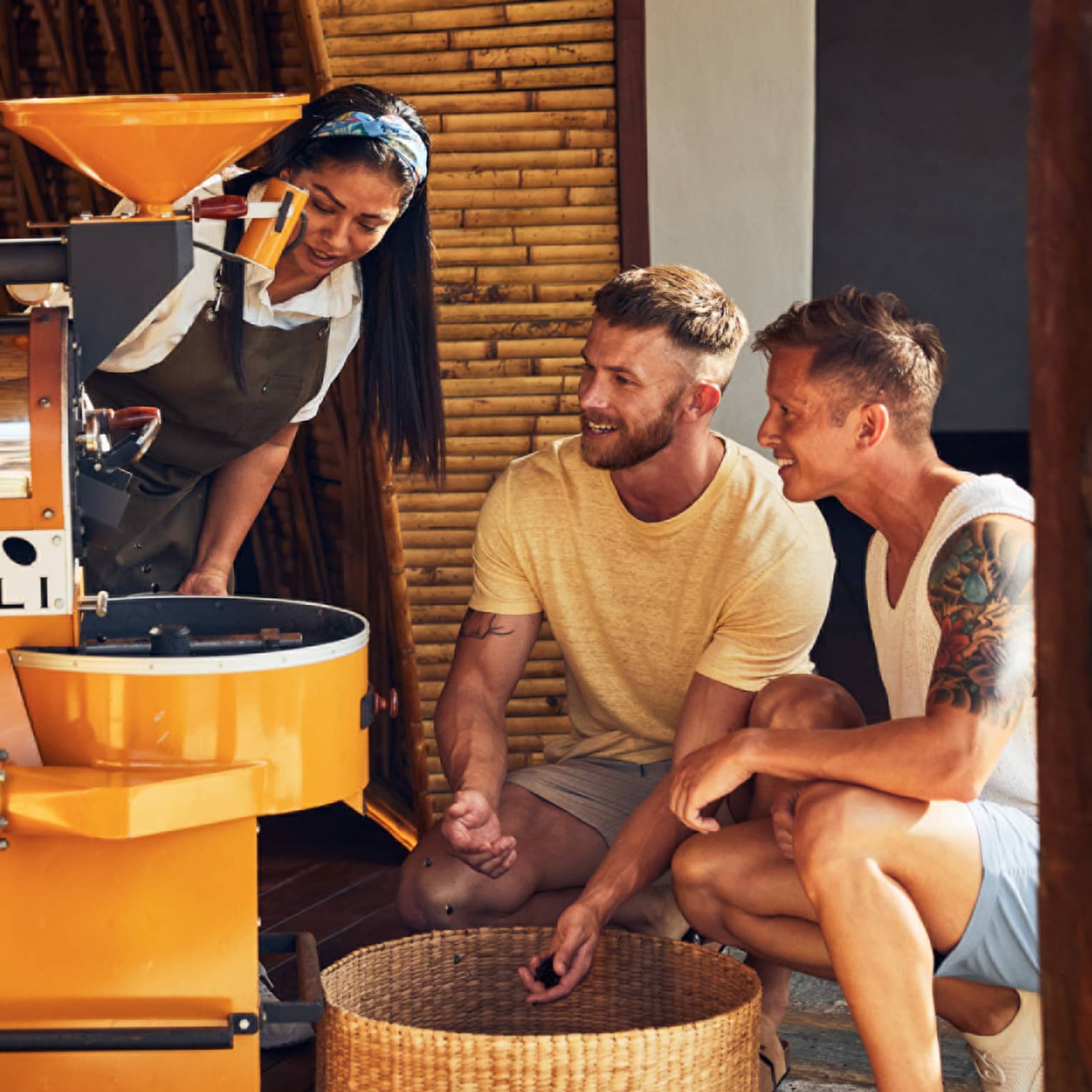 A woman demonstrates coffee roasting to two guests, who observe closely near a yellow coffee roaster in a bamboo-walled setting.