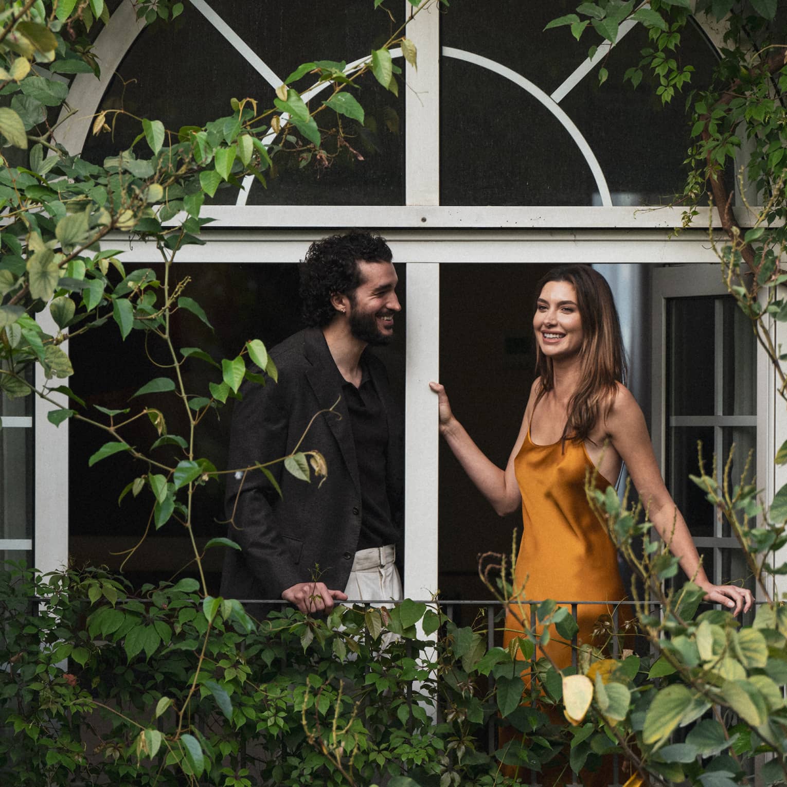 Two people stand on a vine-covered balcony surrounded by foliage.
