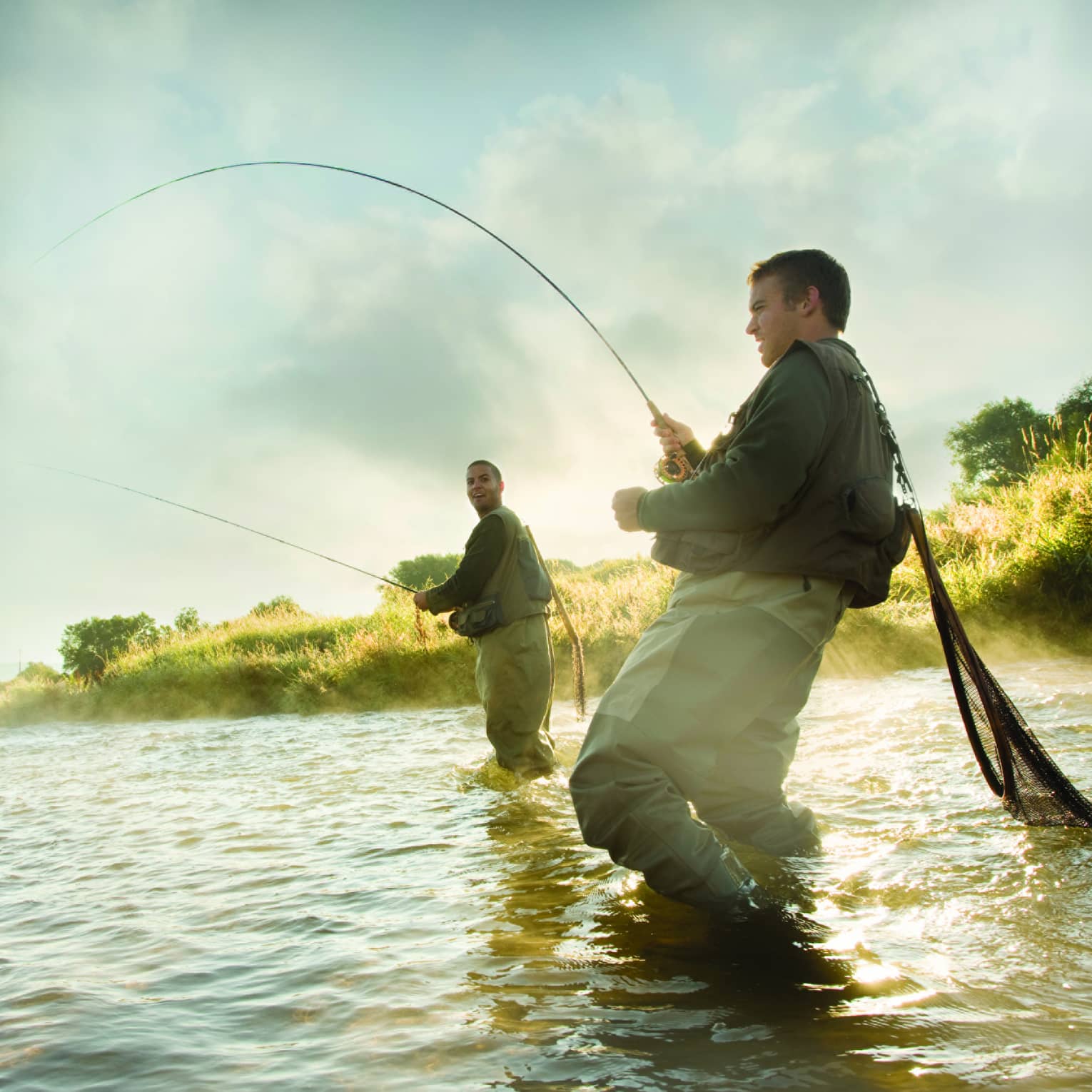 Two people fly fishing in a river, standing knee-deep in the water, surrounded by mist and greenery.