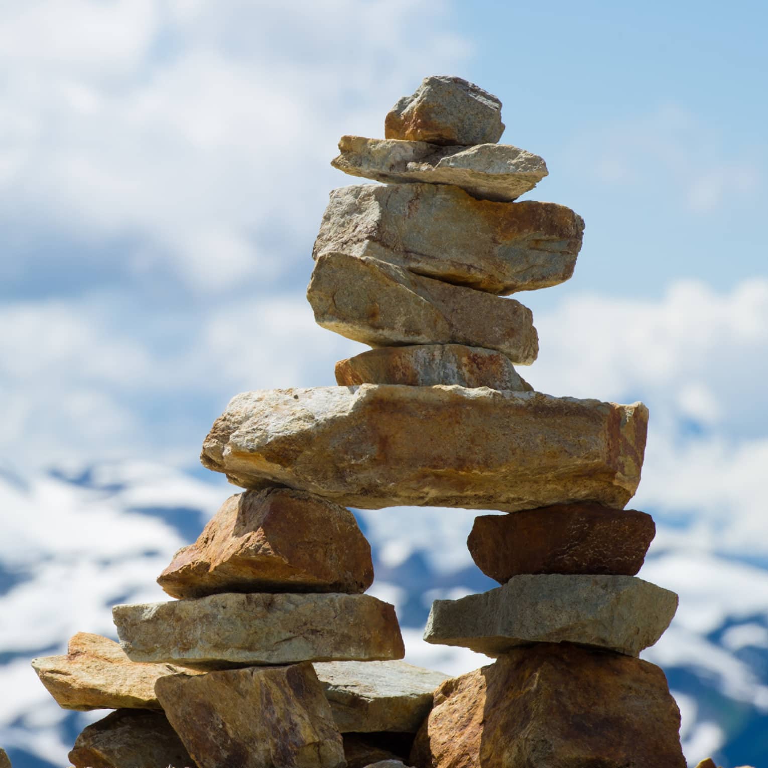 Close-up of an inukshuk atop a high rock shelf, against blue sky, white clouds and a majestic distant mountain.