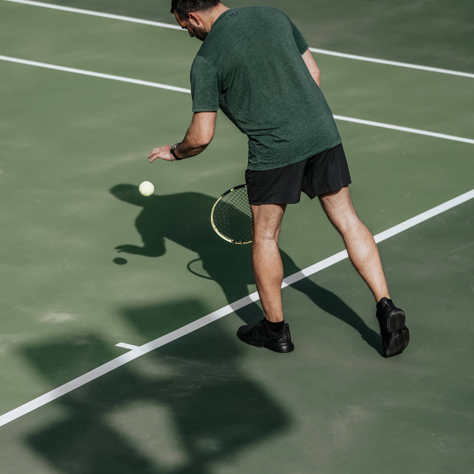 Man on tennis court bouncing tennis ball with left hand, preparing to serve
