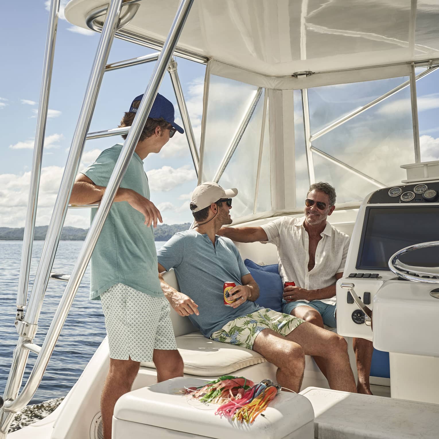 Aboard a fishing vessel on calm water, three people relax under the shaded cockpit, their fishing rods stored beside them.