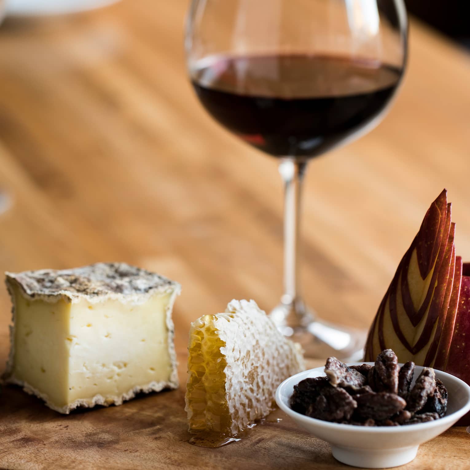 A glass of red wine with a luxurious wooden tray holding cheese, honeycomb, honey, sliced apple, nuts and crackers.