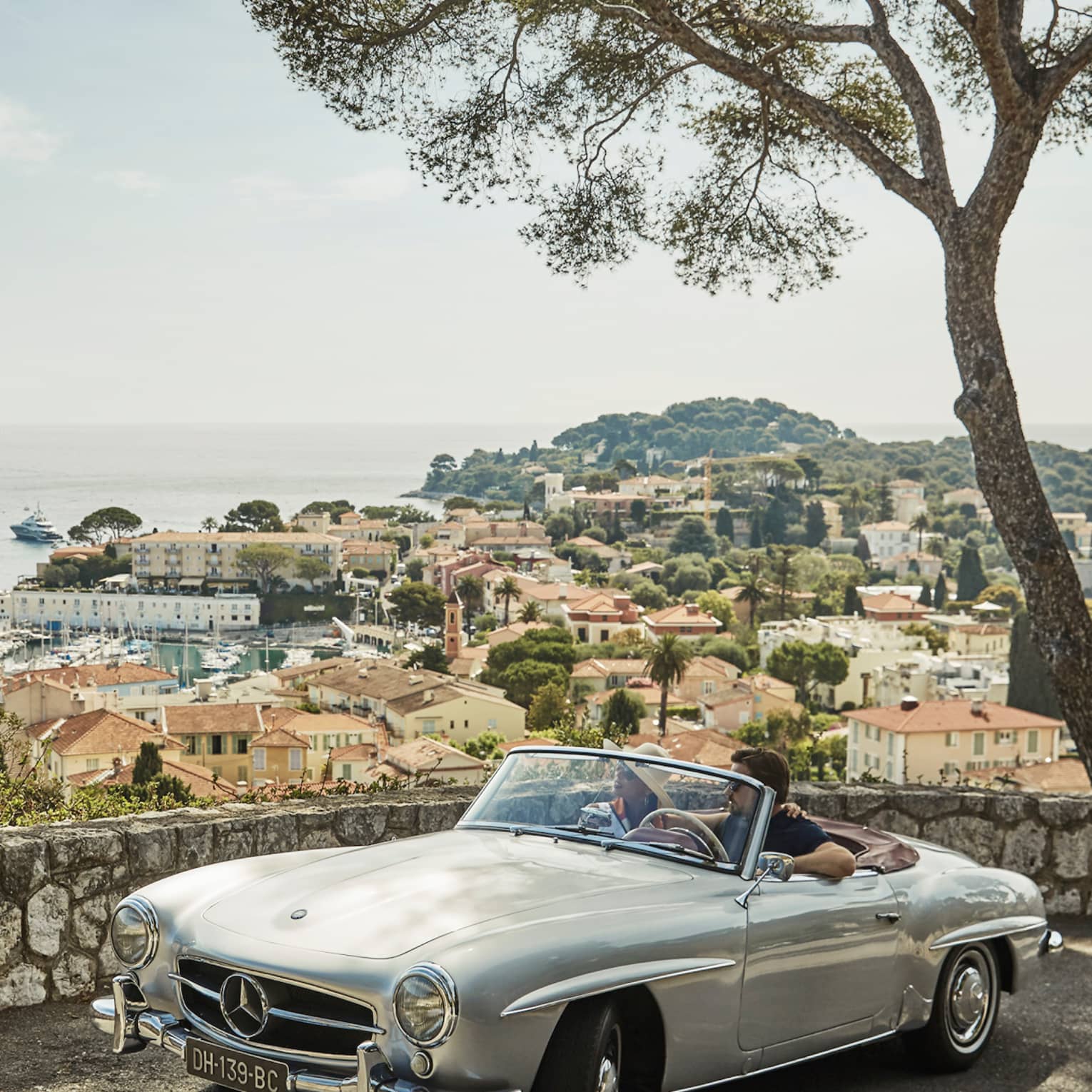 Couple in luxury vintage white car parked under tree by stone wall overlooking red roofs, water