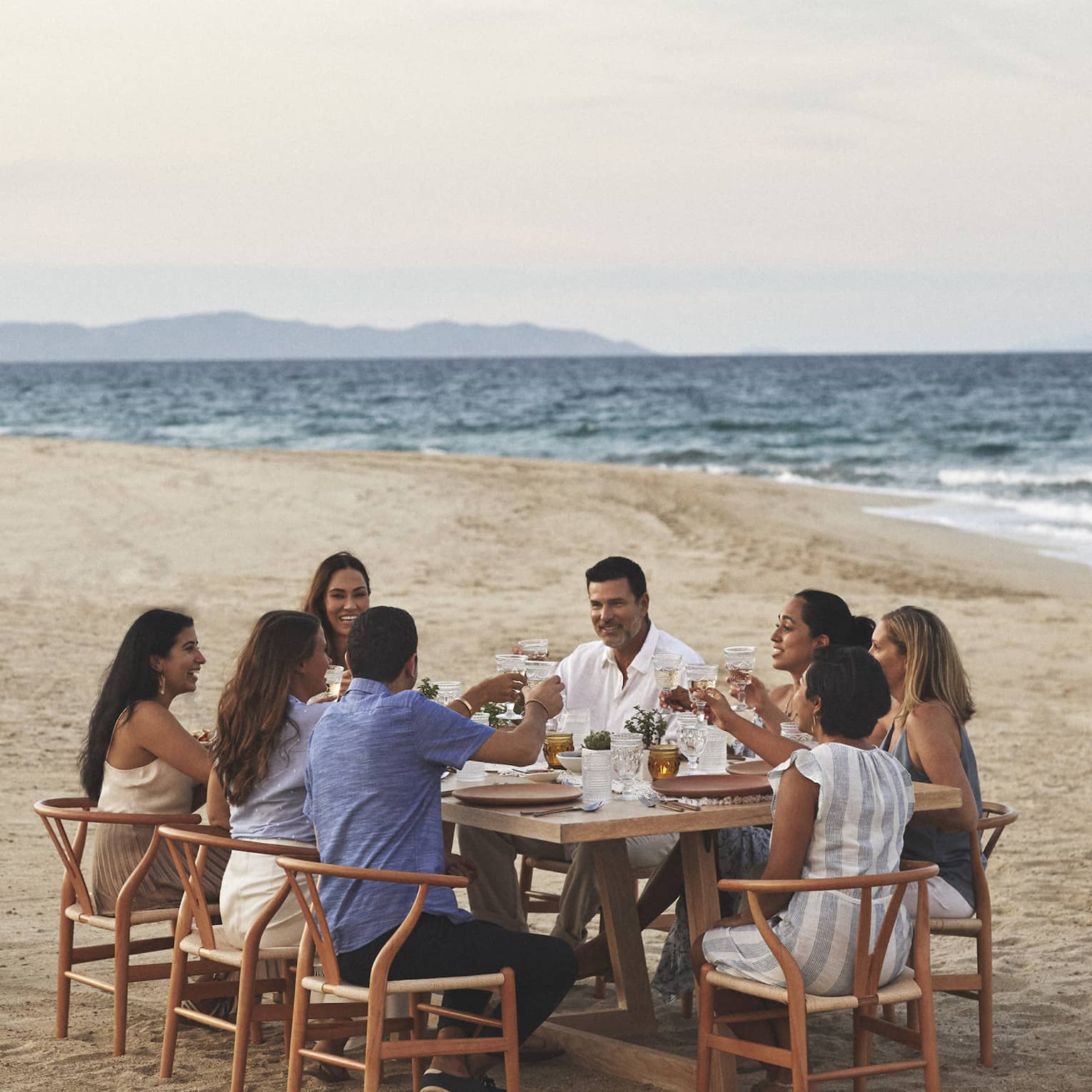 A group dines on the beach in wooden mid century modern dining chairs pulled up around a rectangular wooden dining table