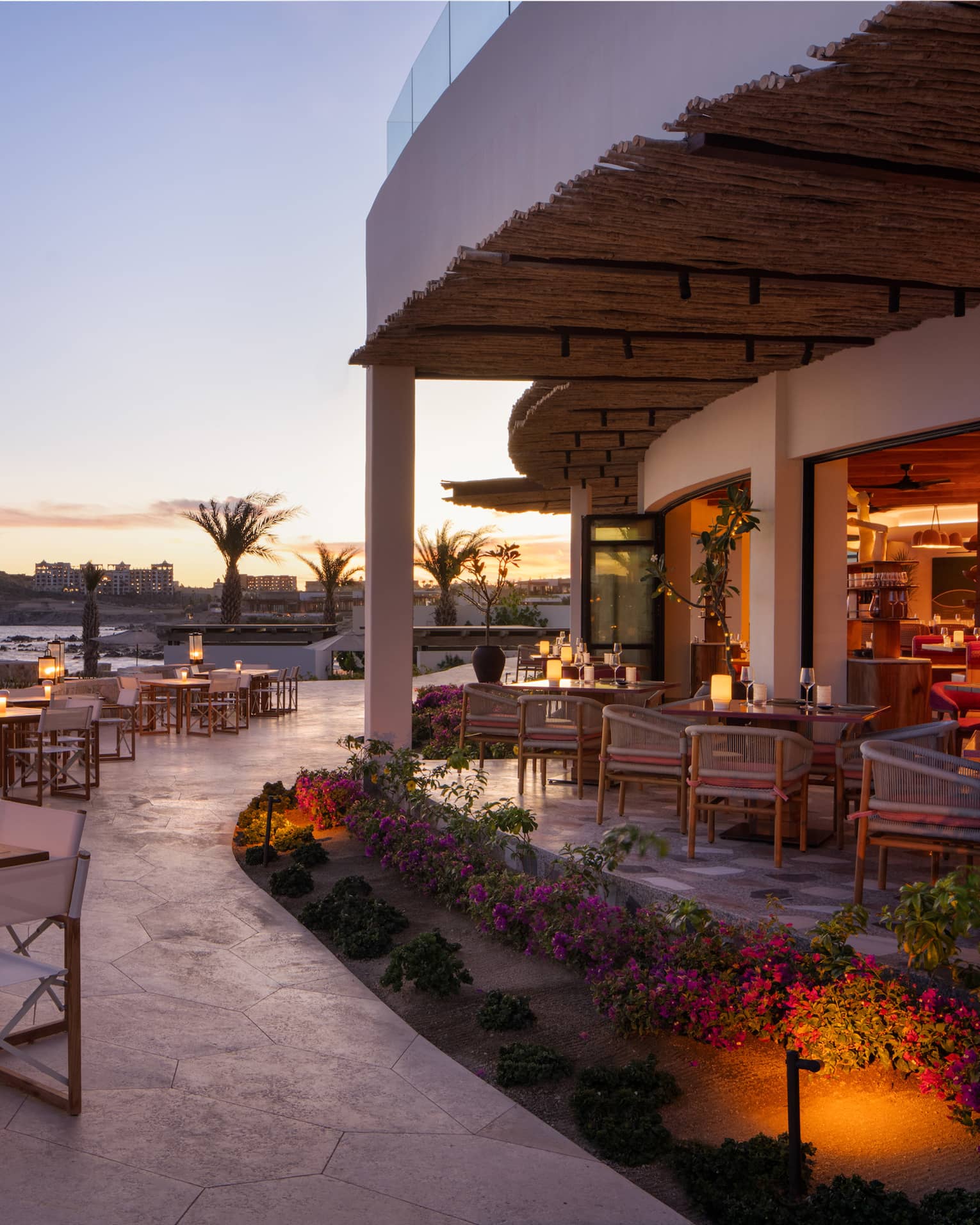 In the warm afterglow of sunset, lanterns light up a large, curved stone restaurant patio at the edge of the ocean.
