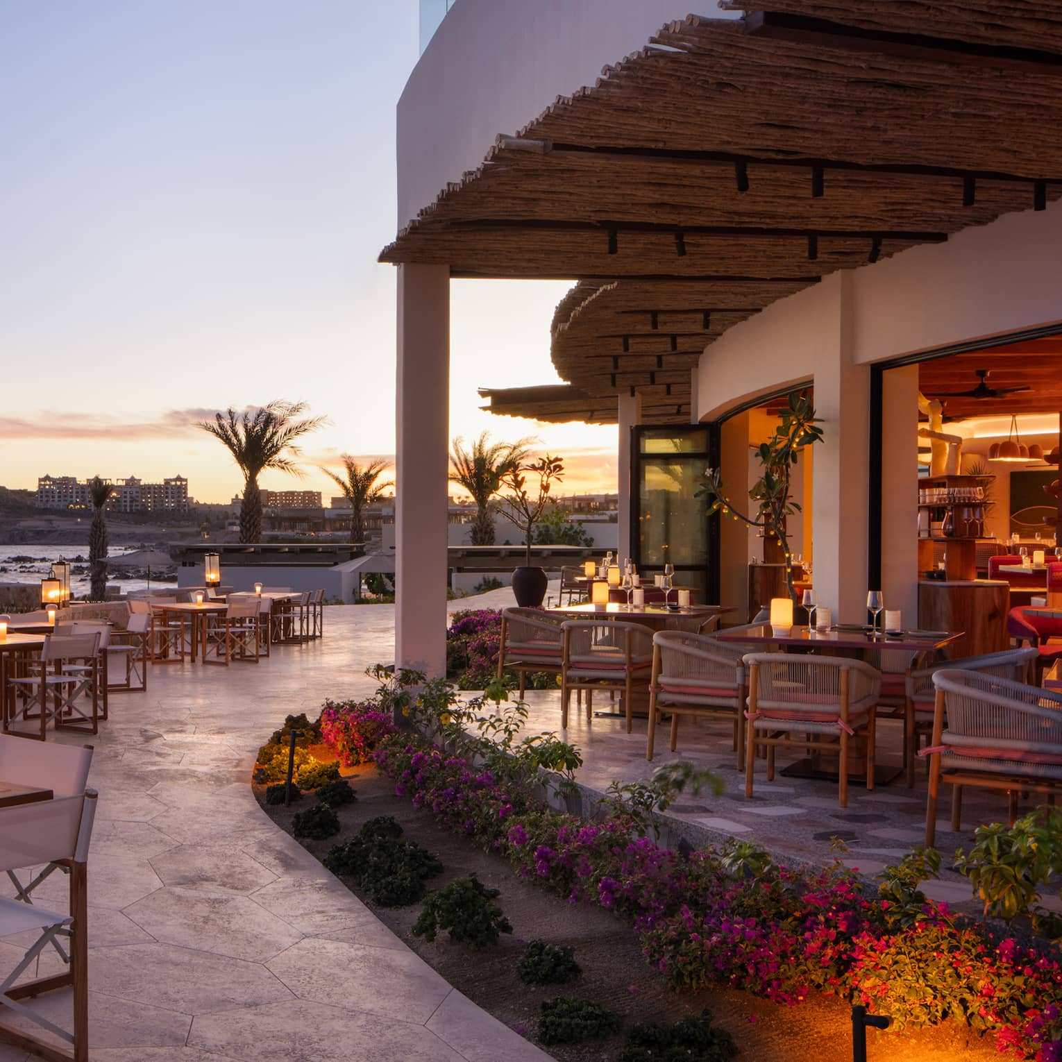 In the warm afterglow of sunset, lanterns light up a large, curved stone restaurant patio at the edge of the ocean.