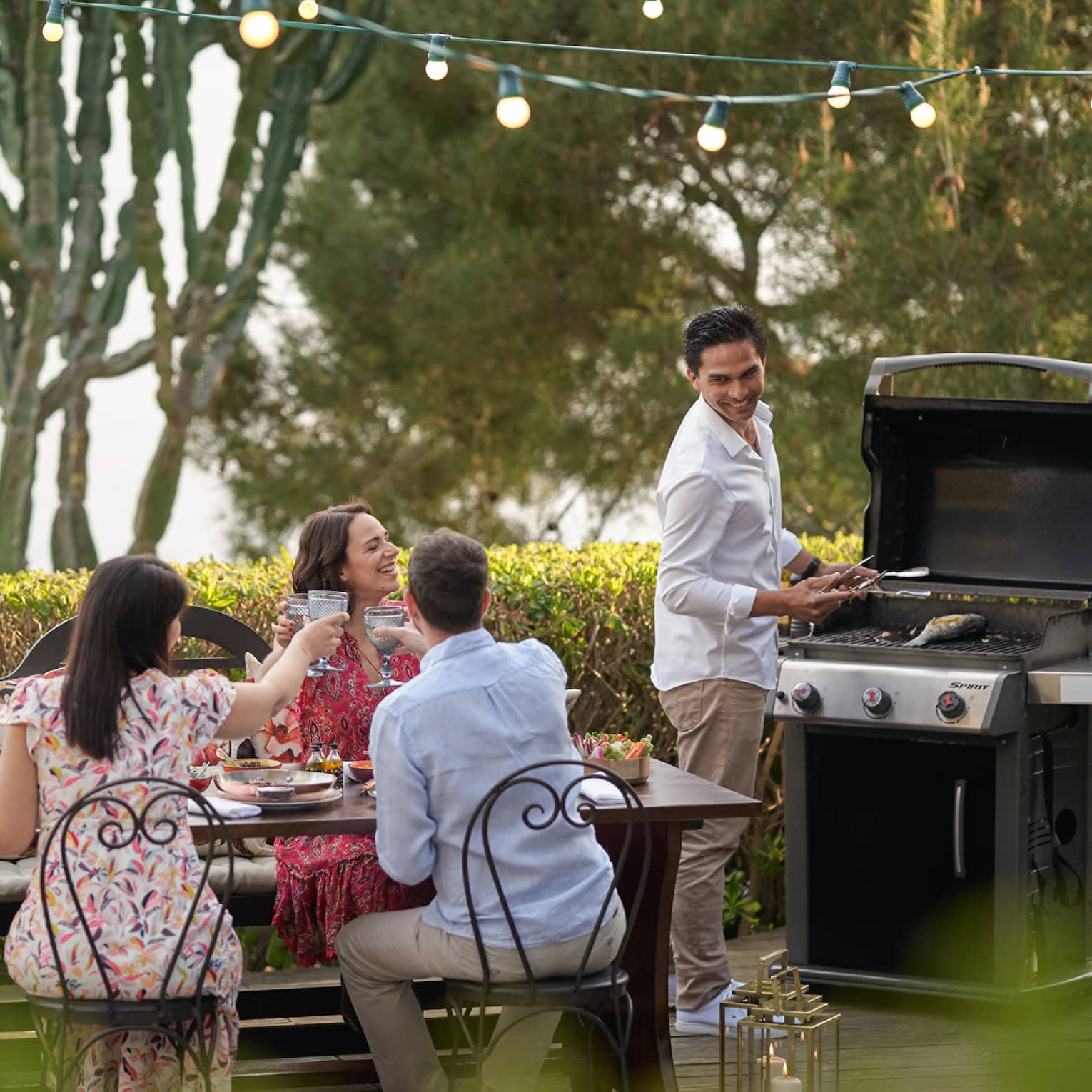 A group of four people grilling food outside at a large table while wearing nice beach clothes.