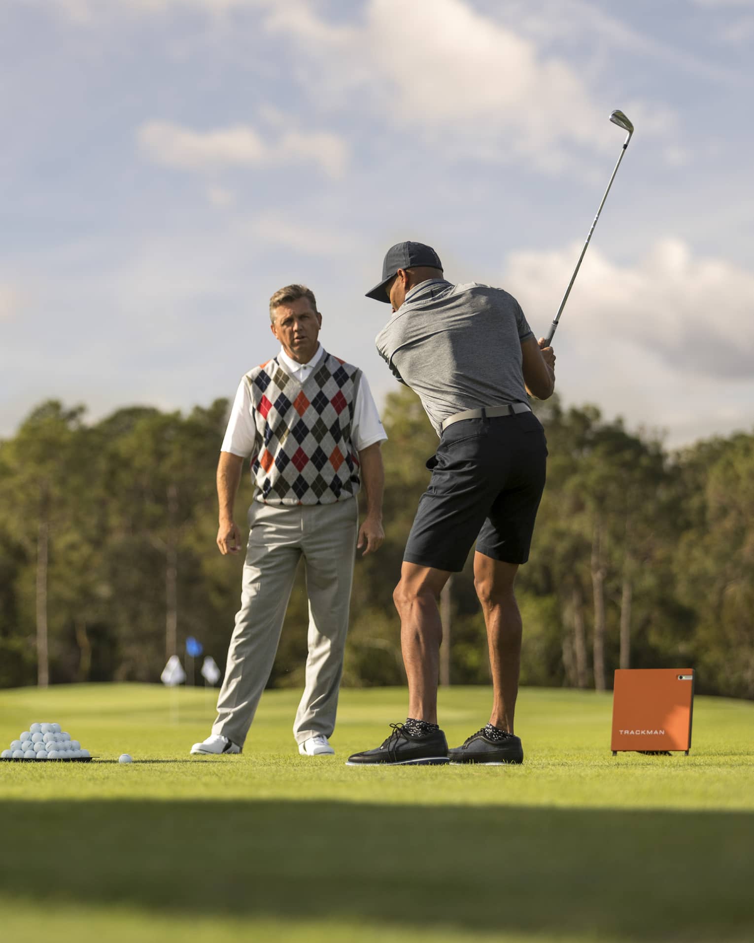 Against clear sky, one golfer looks on as another prepares to swing, a tray of balls and a stand of clubs nearby.