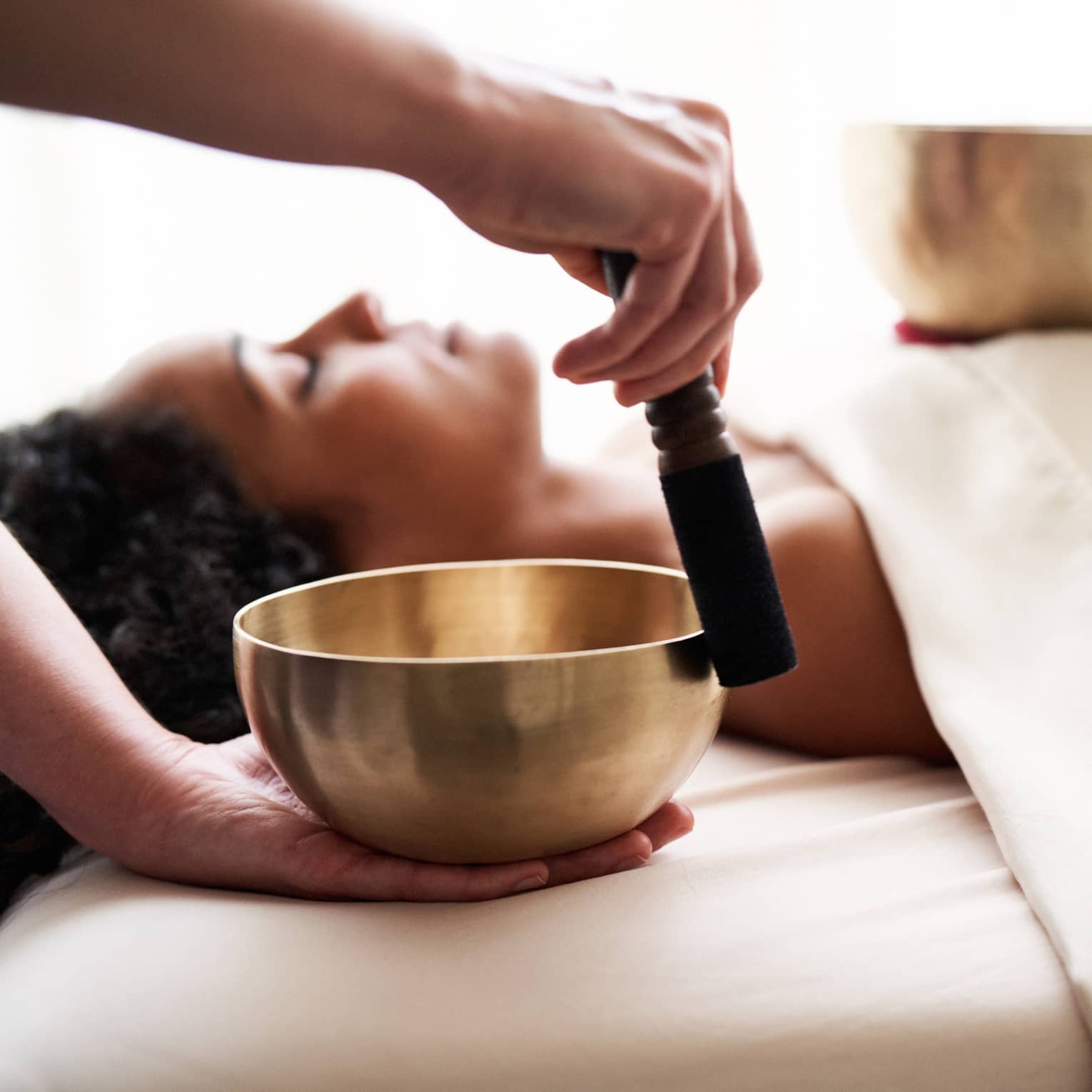 Woman with eyes closed lies on massage table in as vibrational sound therapy is performed with a Tibetan singing bowl