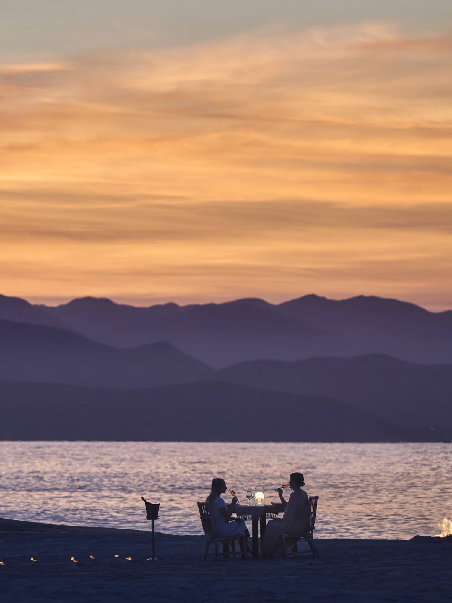 A couple dining on a beach during sunset, the mountains in the background look purple while the sky is orange and pink.