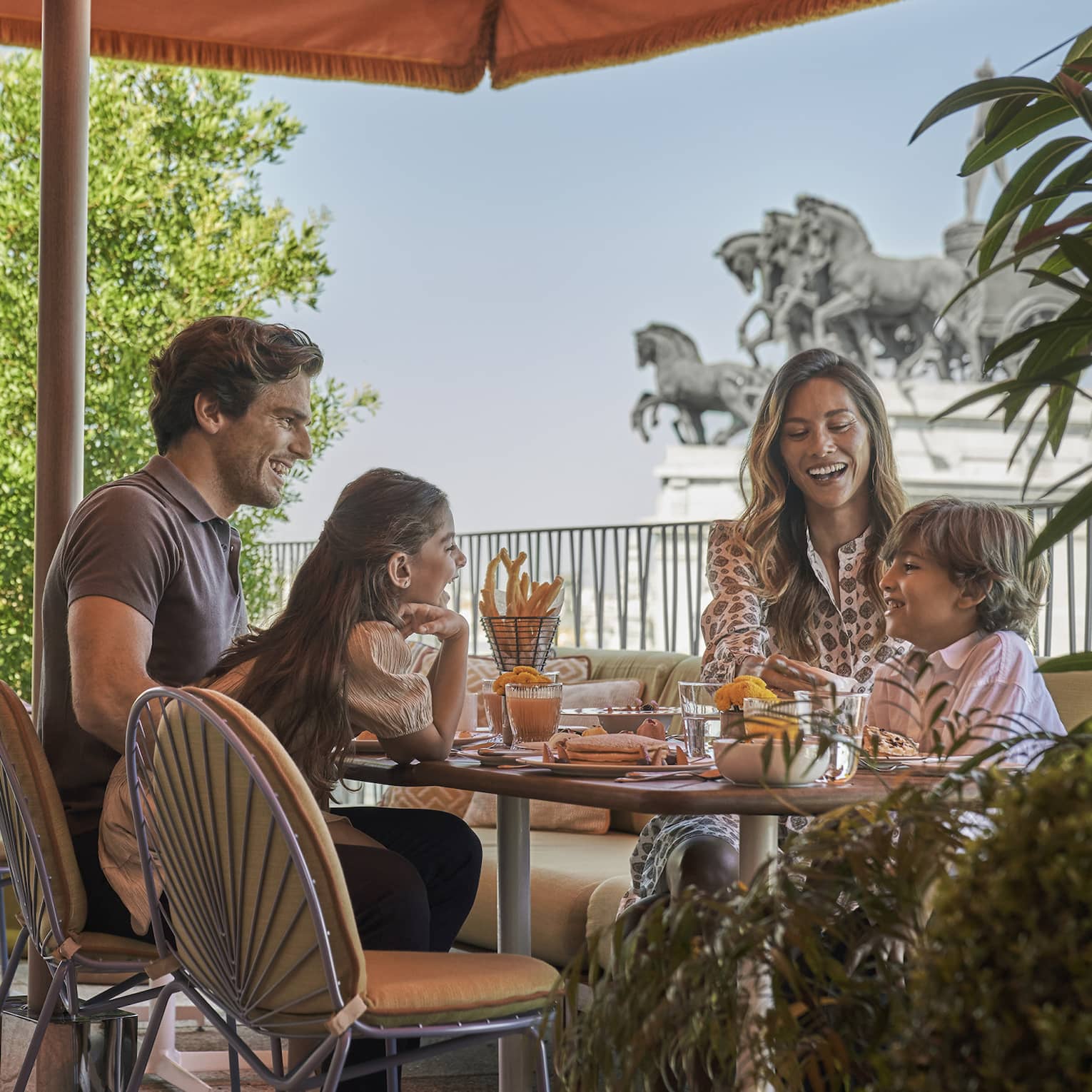 Family enjoying breakfast outdoors under an orange canopy, with views of horse statues in the background