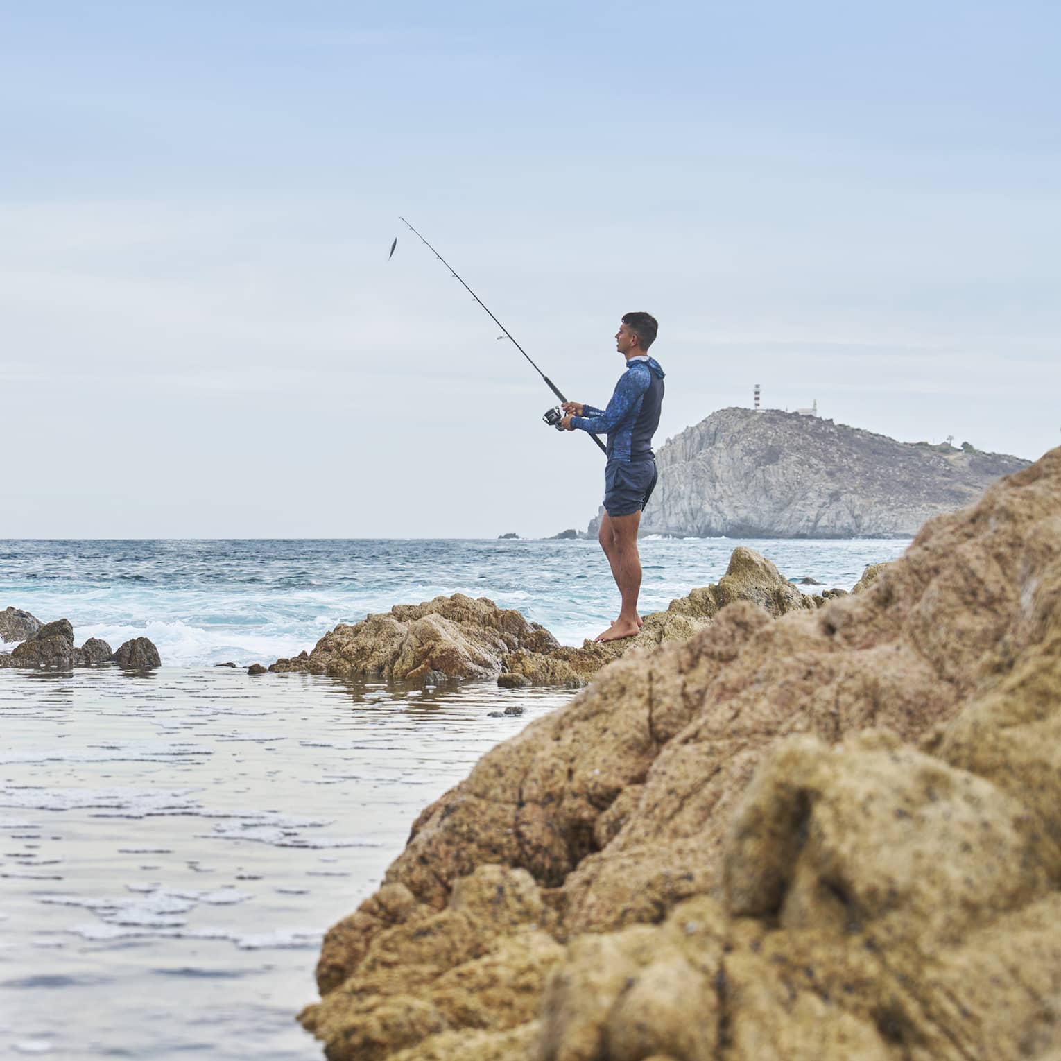 A fisher holding a pole stands barefoot atop a rocky outcrop and looks out toward an expanse of blue water under a clear sky.