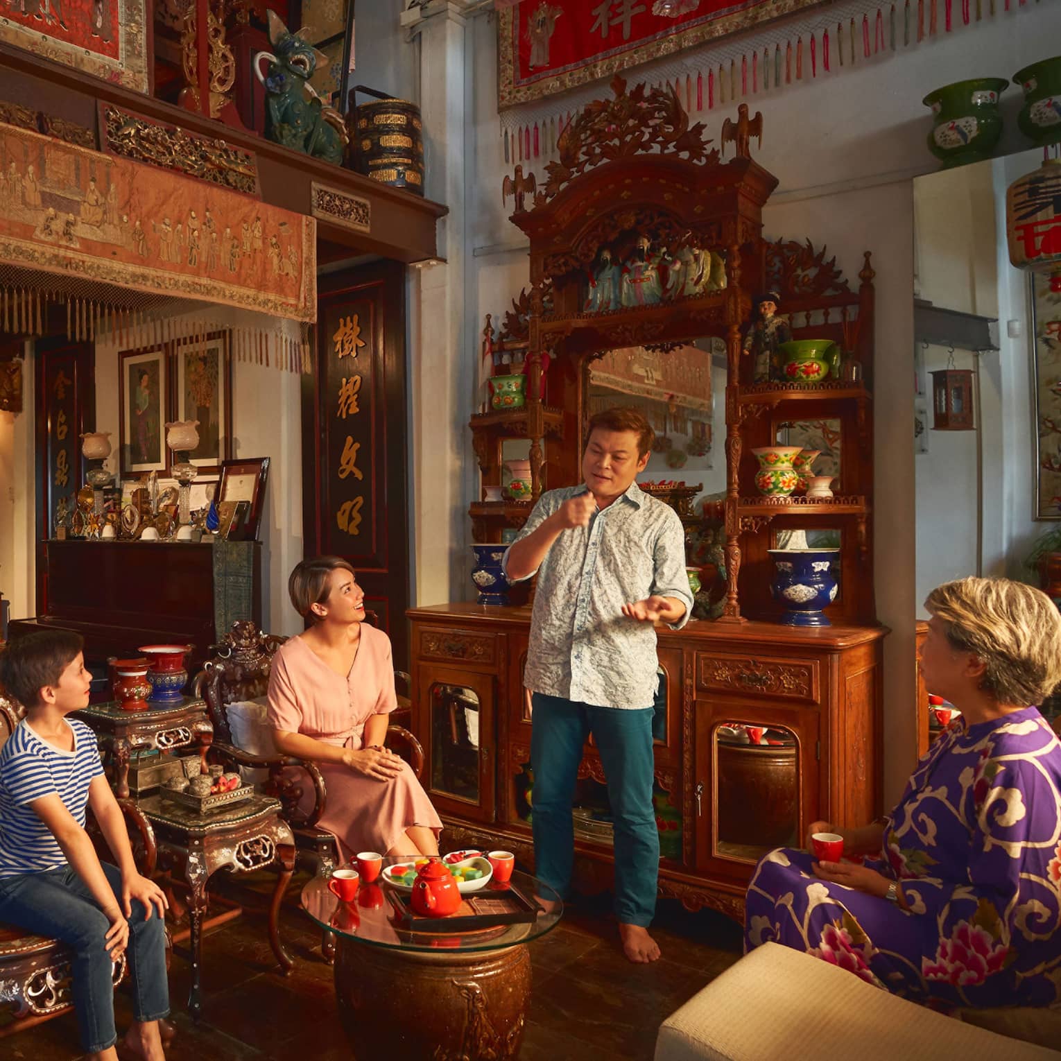 A family gathers around a low, round table holding a tea set in a room with ornate furniture, porcelain vases and tapestries.