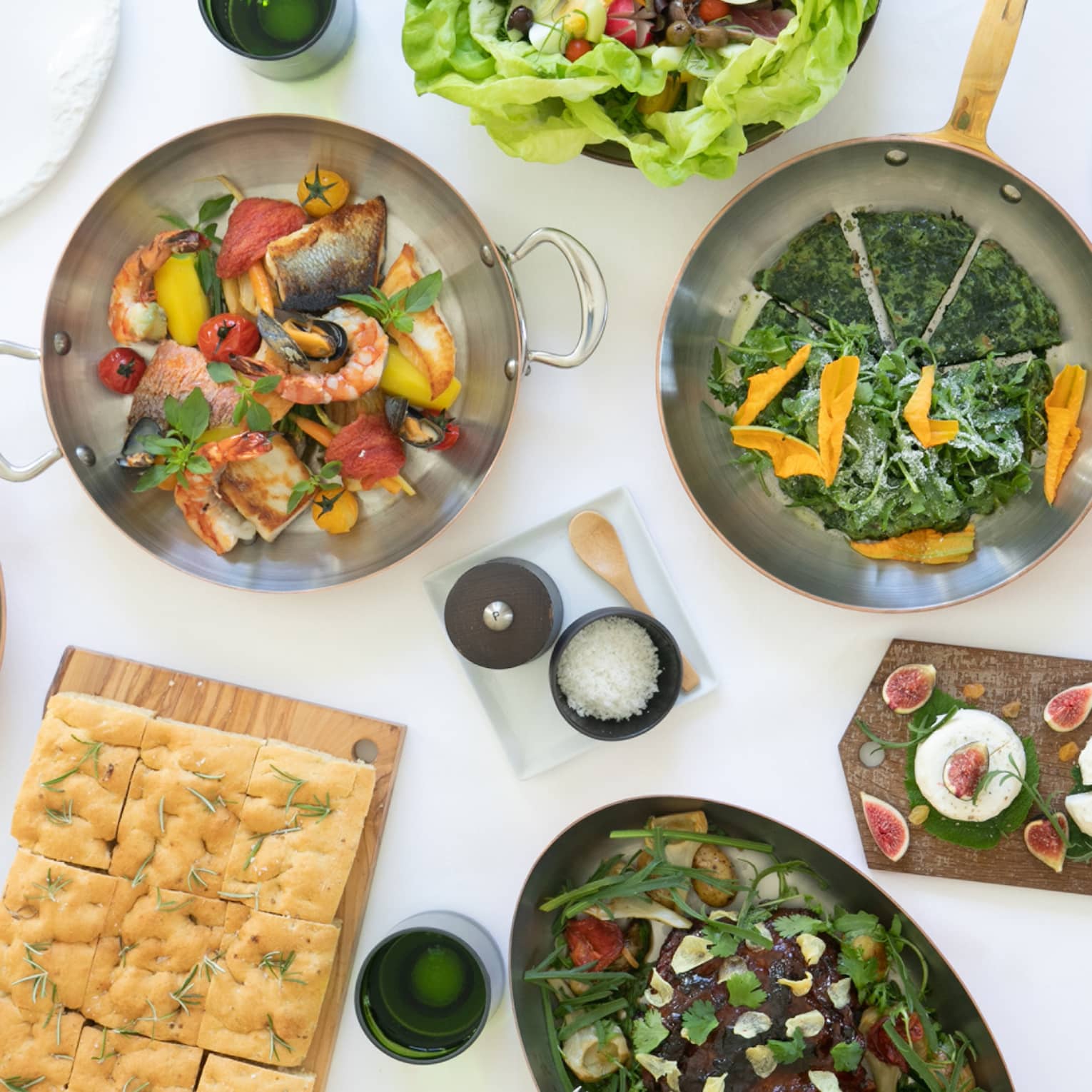 View from above a stark white surface covered in dishes, trays and pans of food, including salads, bread, figs and seafood.