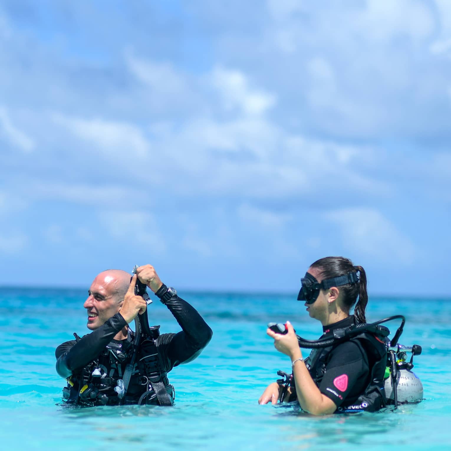 Scuba instructor showing mouthpiece to guest as they stand wearing scuba gear, chest-deep in azure water under fluffy clouds.