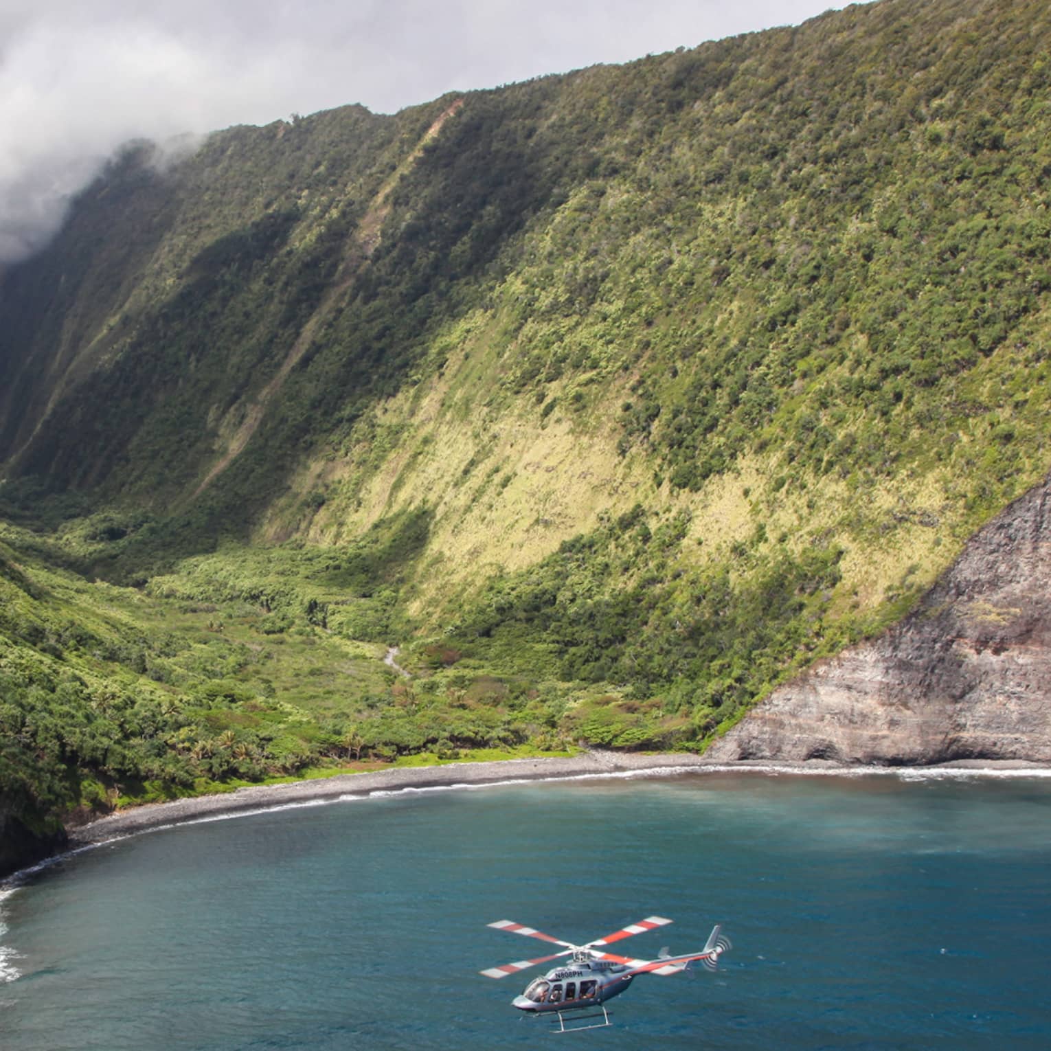 Helicopter flies over ocean by sweeping green volcanic mountains