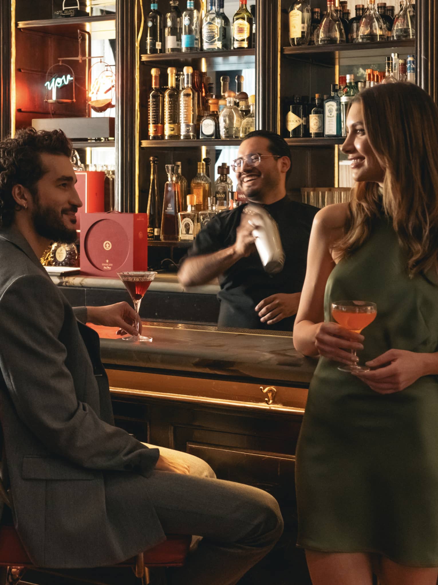 People socialize at a bar counter; a bartender making a drink in a shaker is in the background.
