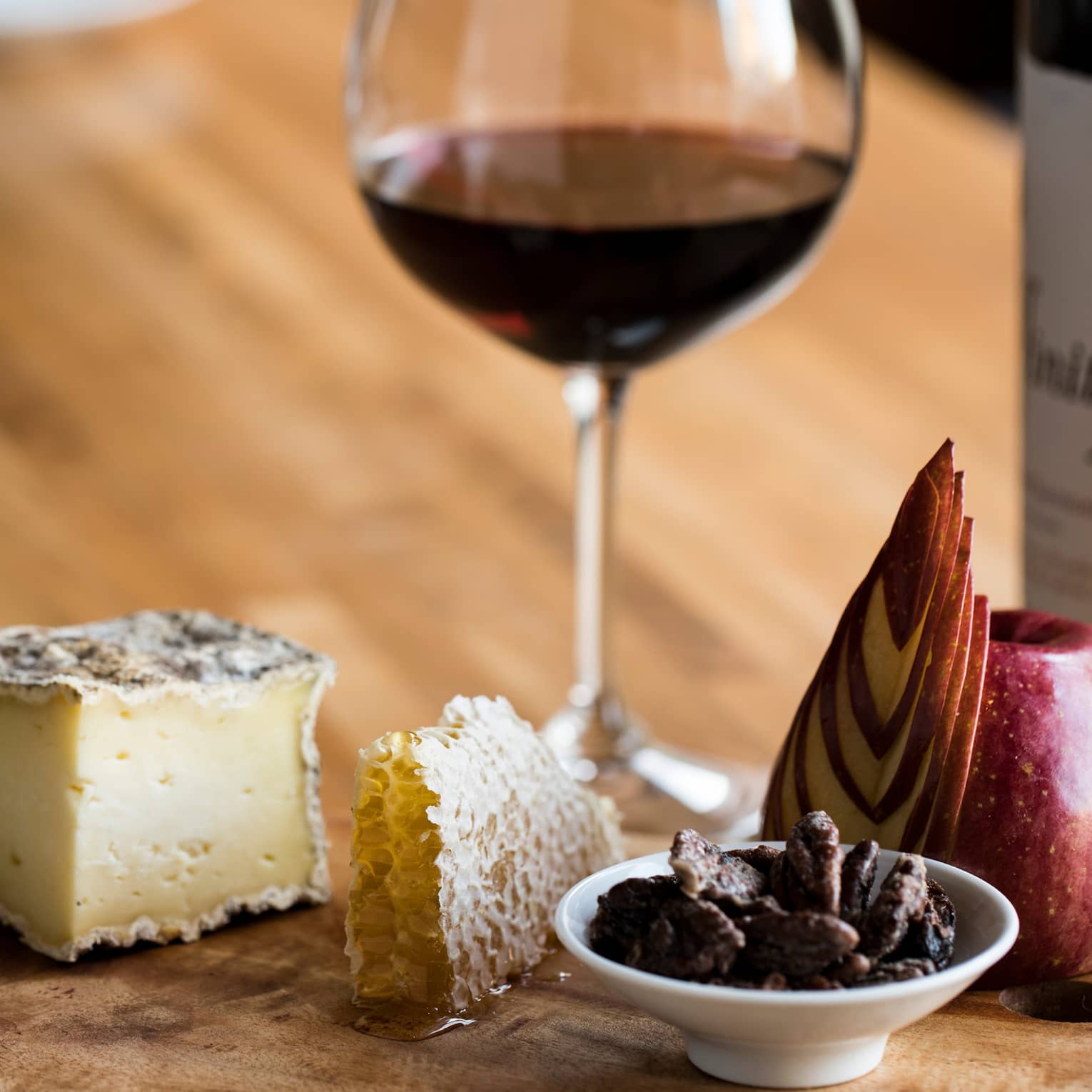 A glass of red wine with a luxurious wooden tray holding cheese, honeycomb, honey, sliced apple, nuts and crackers.