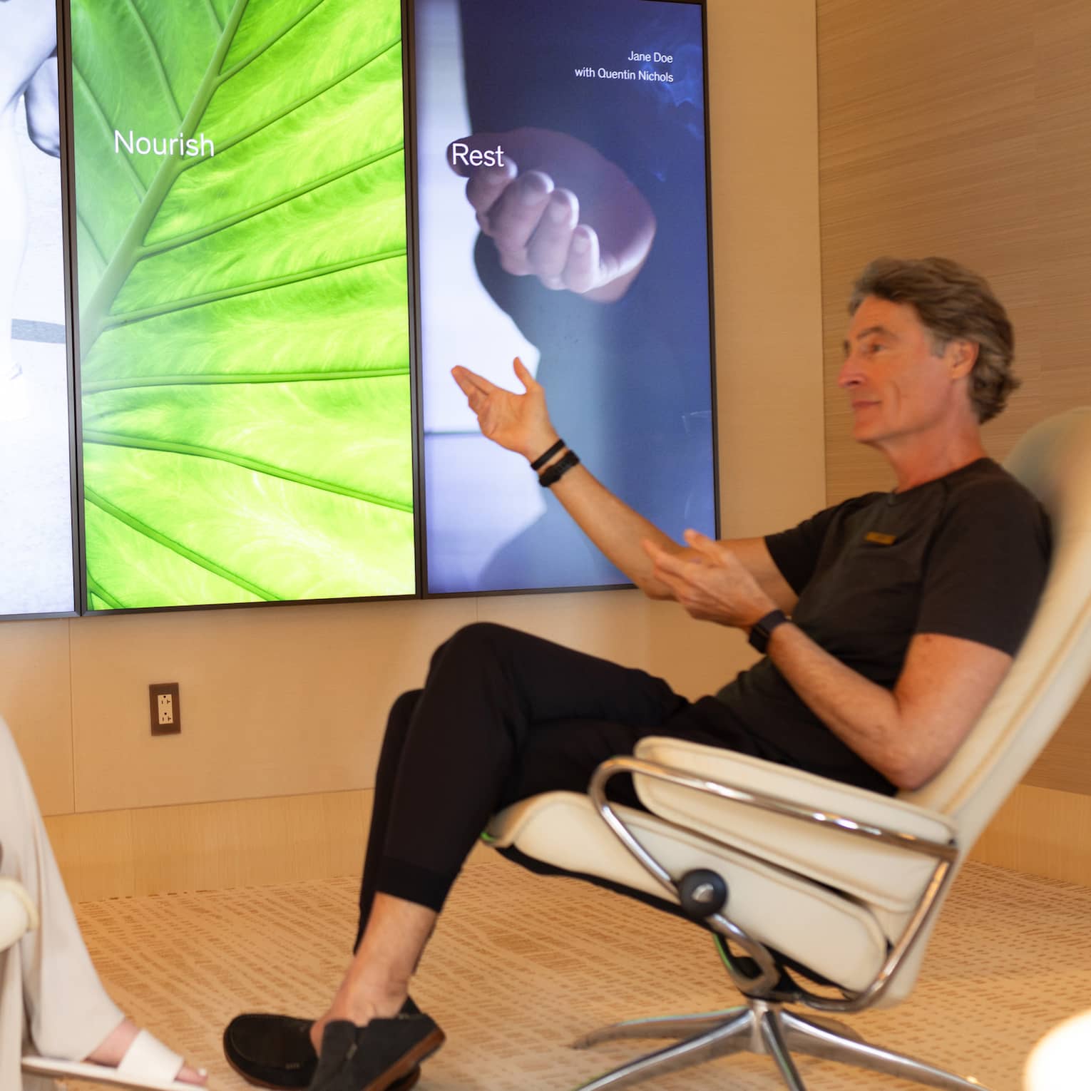 Man and woman sit in arm chairs discussing biomarker results on a TV screen
