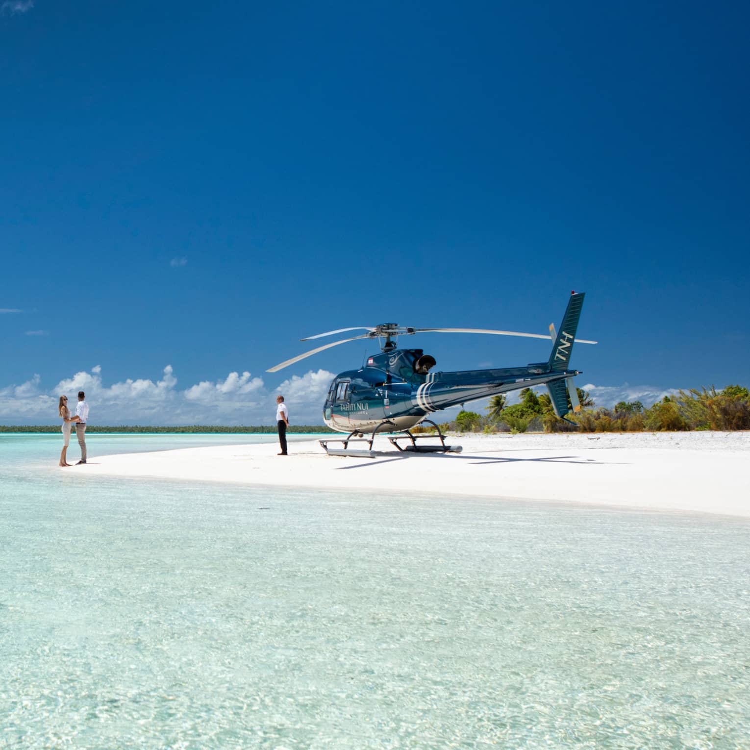 A couple holds hands on a white sand beach beside the pastel blue ocean; a parked helicopter and its pilot wait nearby.